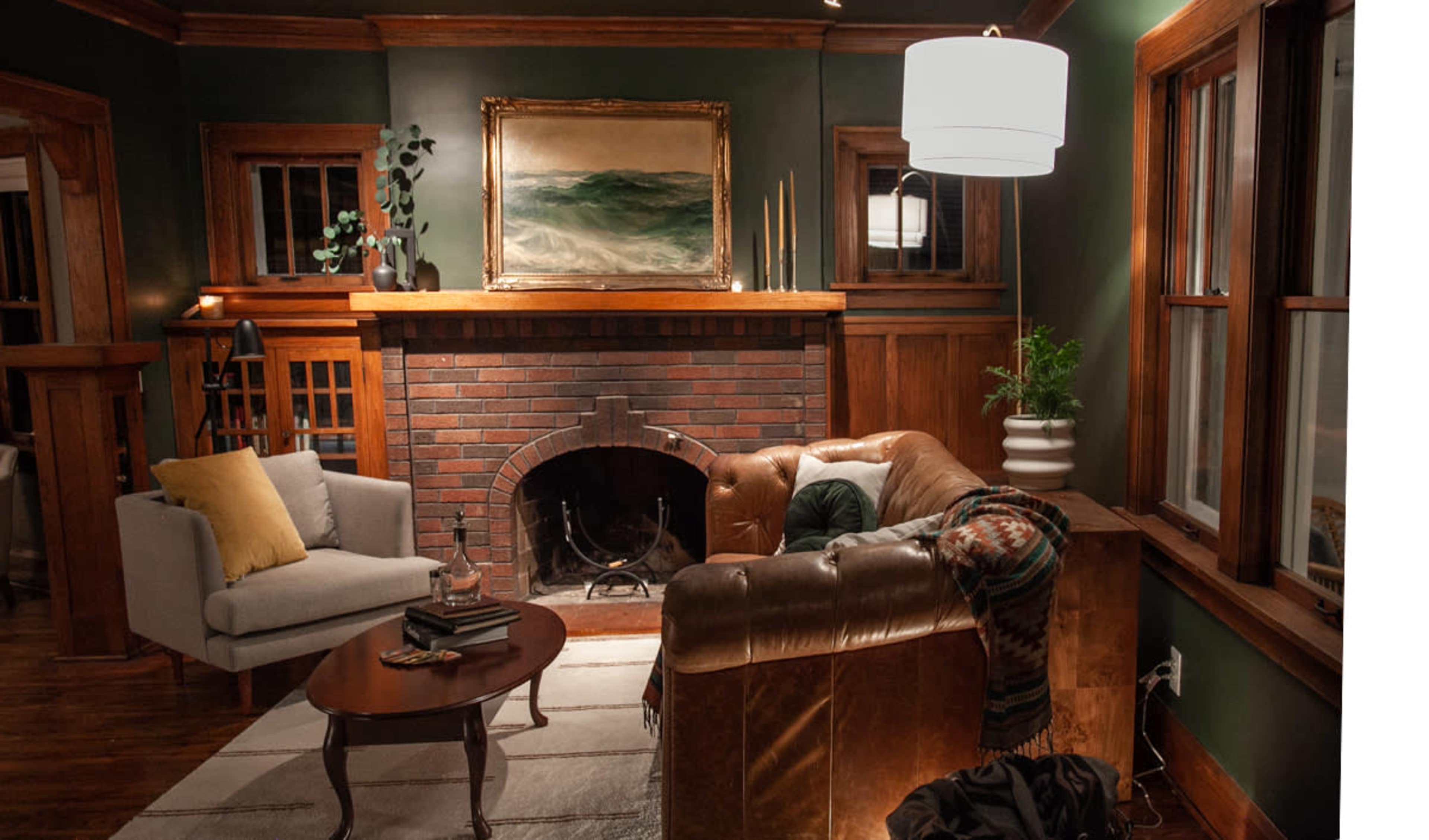 A cozy living room featuring a brown leather sofa, a light gray armchair, a wooden coffee table, and a decorative fireplace framed by dark wood paneling and a large painting above it.