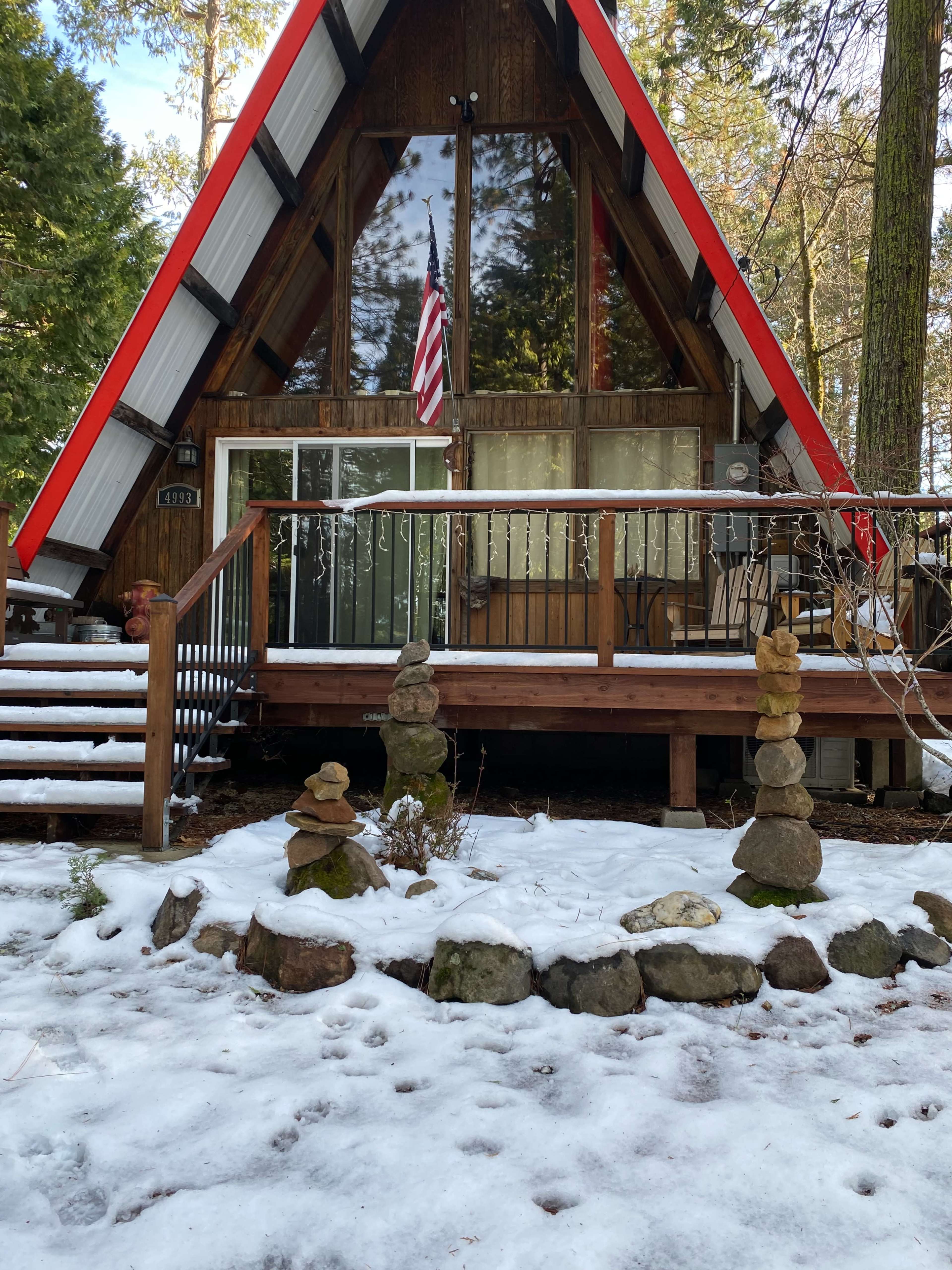 A triangular cabin with a red roof and an American flag is situated in a snowy forest setting, featuring a wooden deck and stacked stone formations in the foreground.