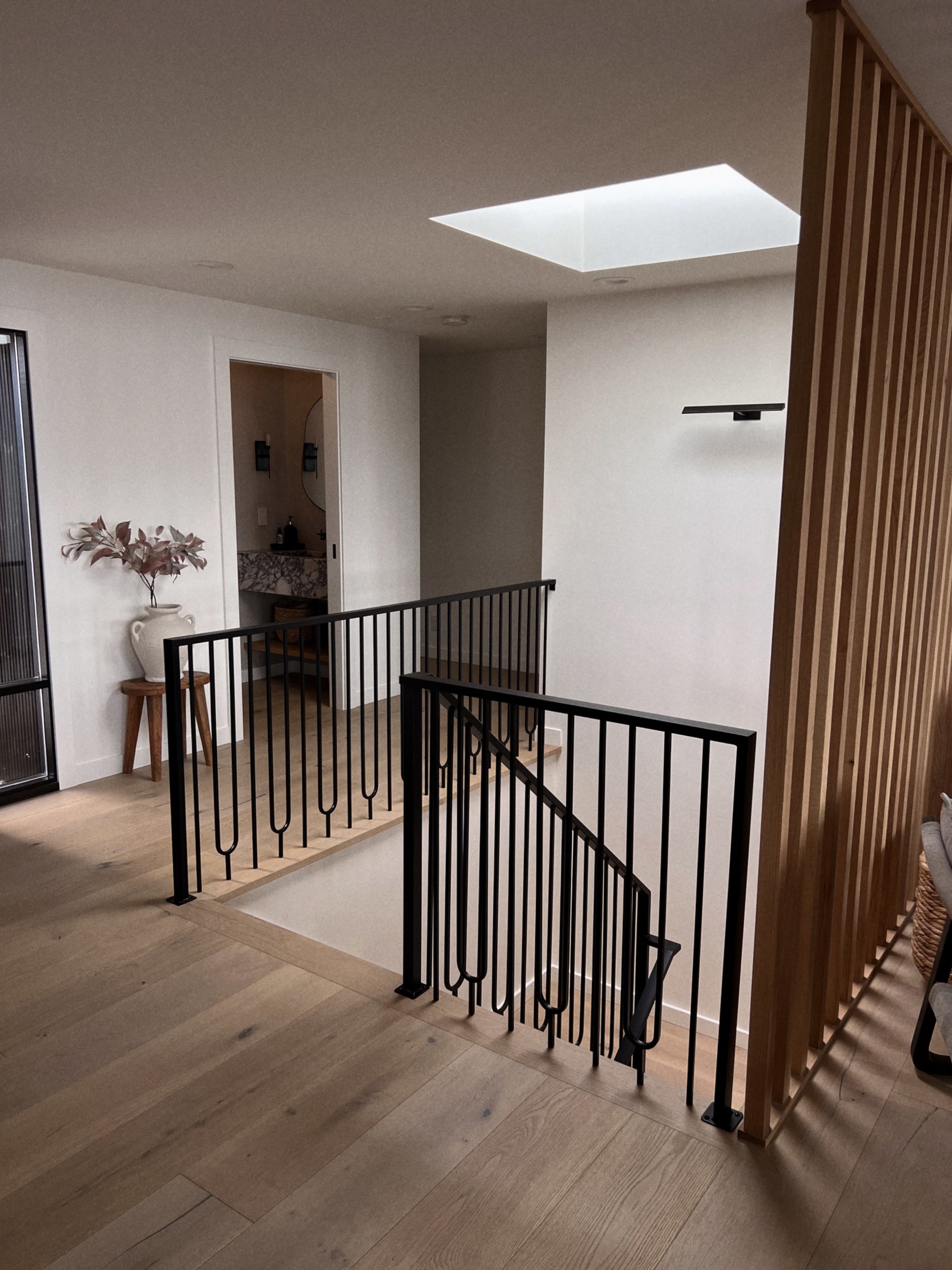 The image shows a modern staircase with a black metal railing, surrounded by light wood flooring and walls, featuring a skylight above and minimal decor.