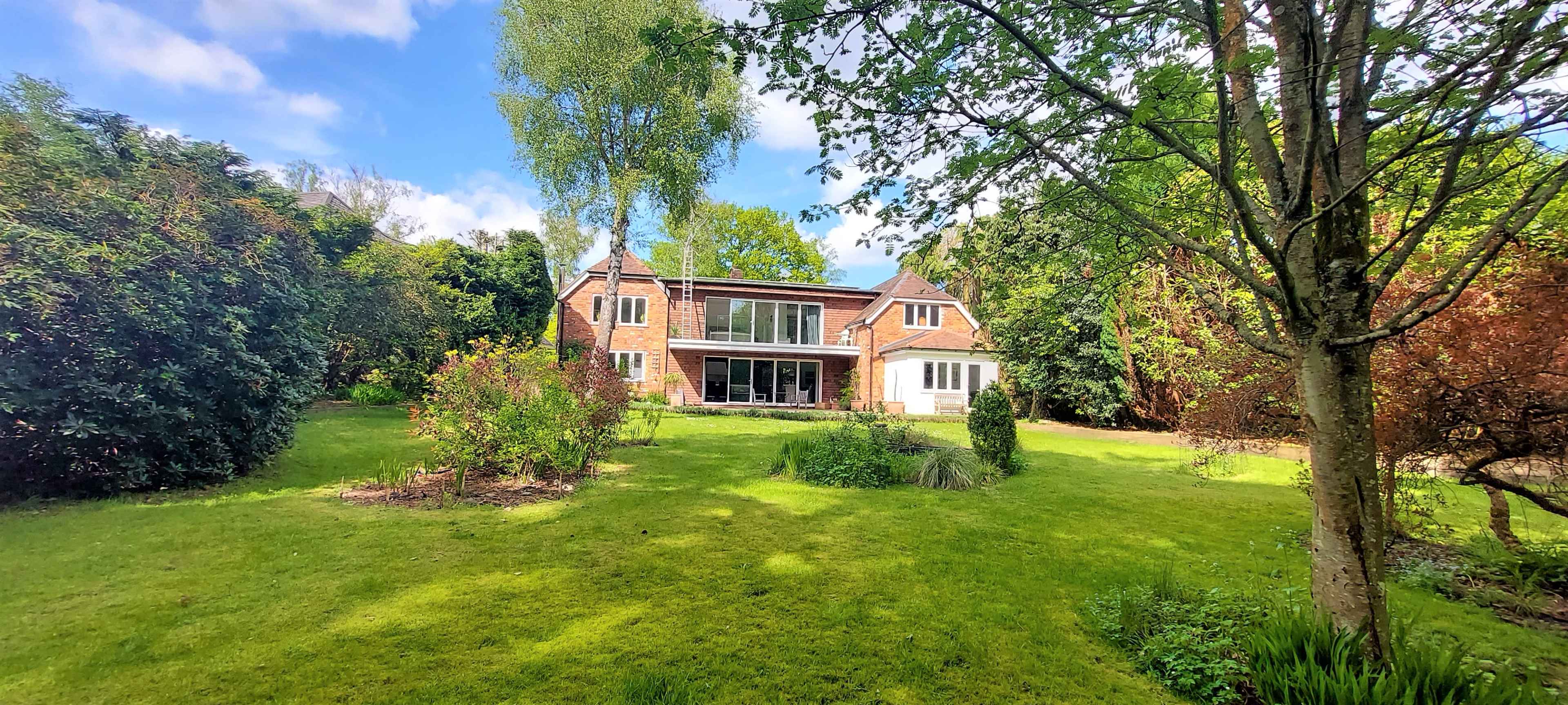A modern two-story house with large windows is set in a lush green garden surrounded by trees and shrubs.