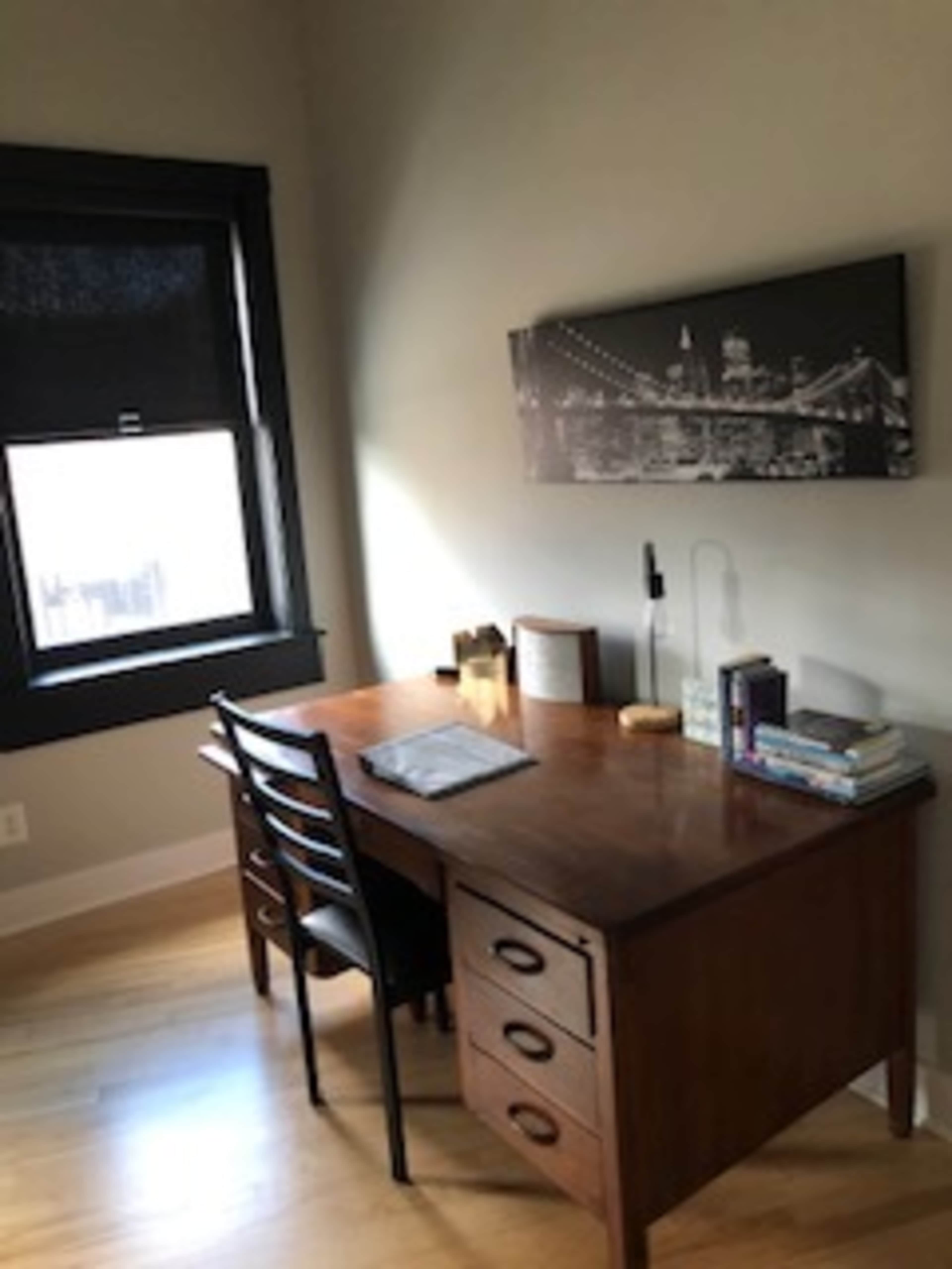 A wooden desk with a chair, books, and a lamp is positioned near a window in a room with light-colored walls.