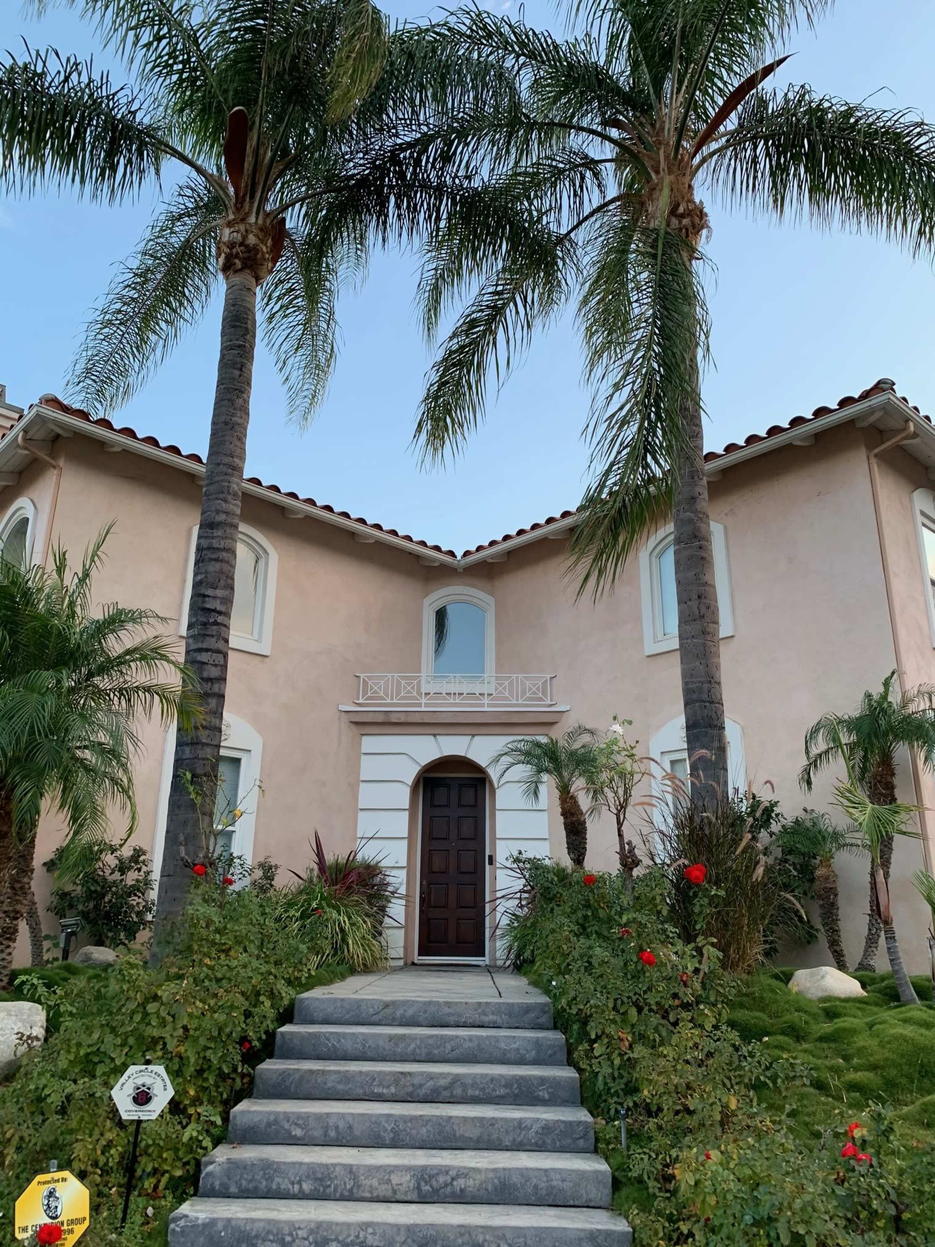 A two-story house with a brown door is flanked by palm trees and surrounded by colorful landscaping.