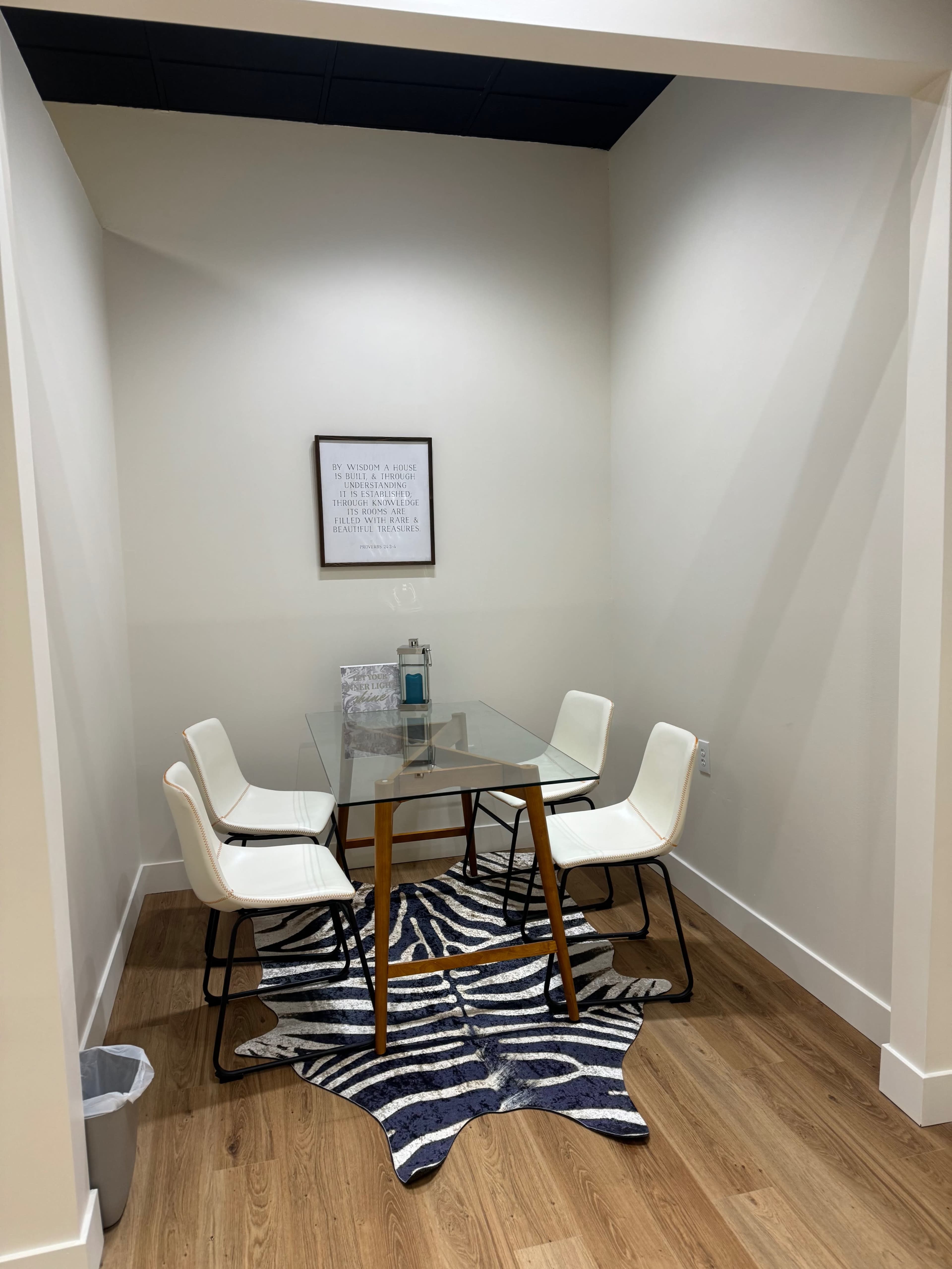 A small meeting space features a glass table surrounded by four white chairs, with a patterned rug underneath and a framed print on the wall.
