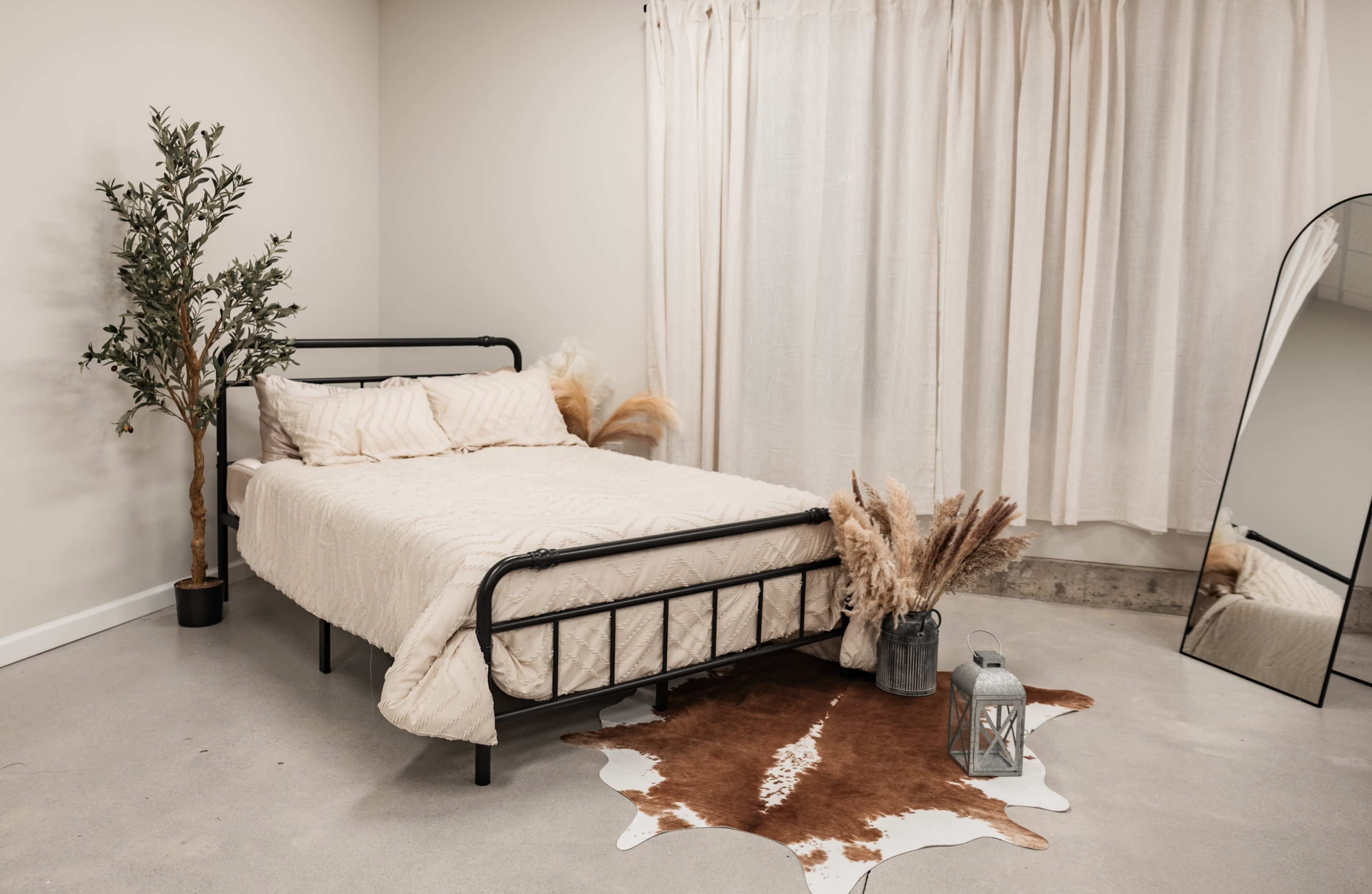 A metal bed with a beige quilt is positioned in a minimalist room featuring a decorative plant, a cowhide rug, and a lantern beside a large mirror.