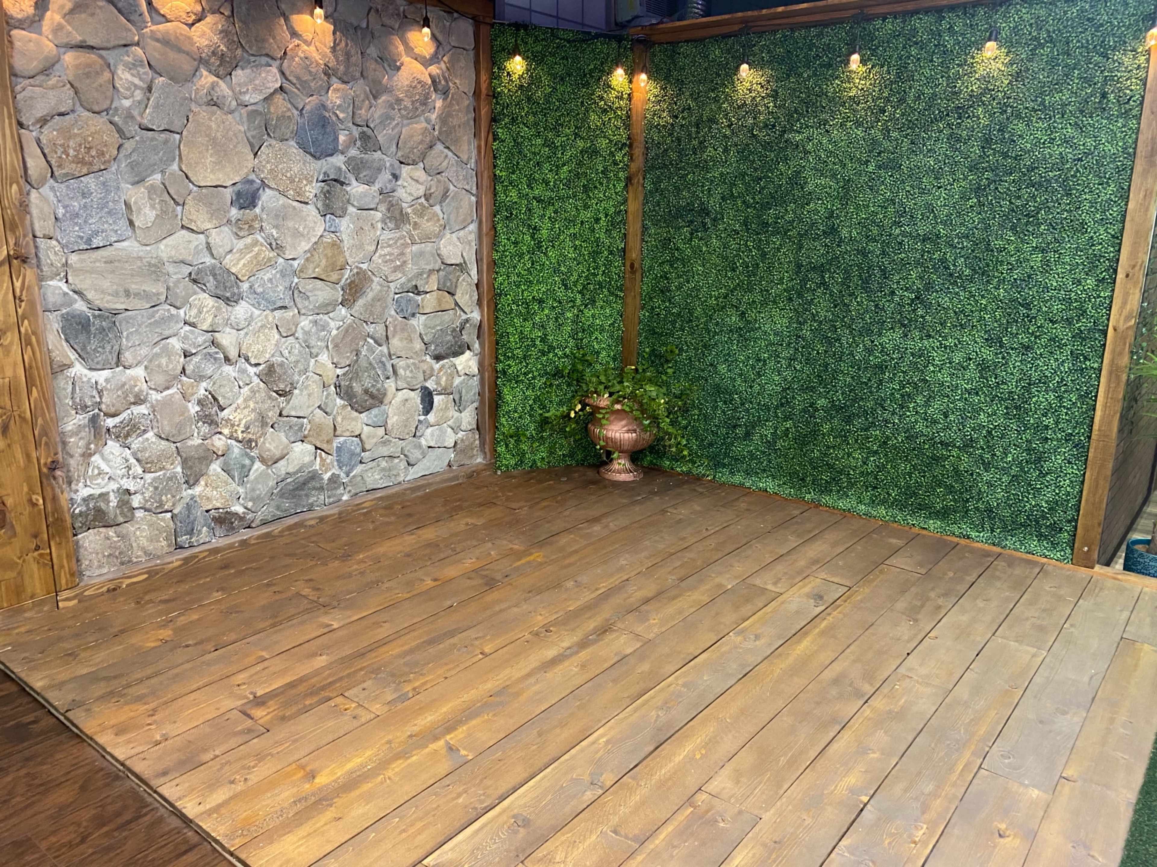 A staged interior space featuring a wooden floor, a stone wall, and a green foliage backdrop with a decorative planter.