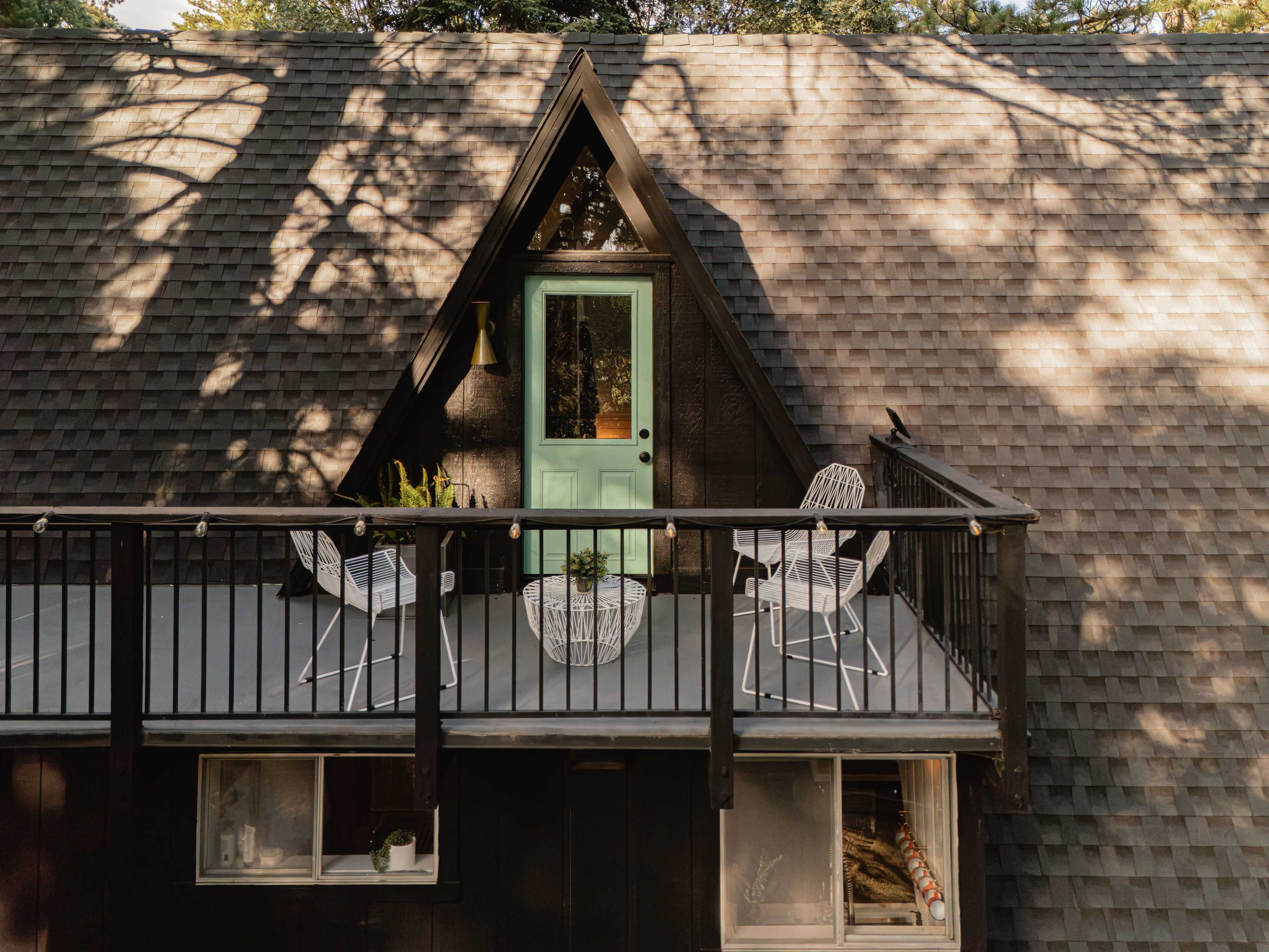 The image shows a balcony with two white chairs and a small table, situated on the upper level of a black A-frame house with a light green door.