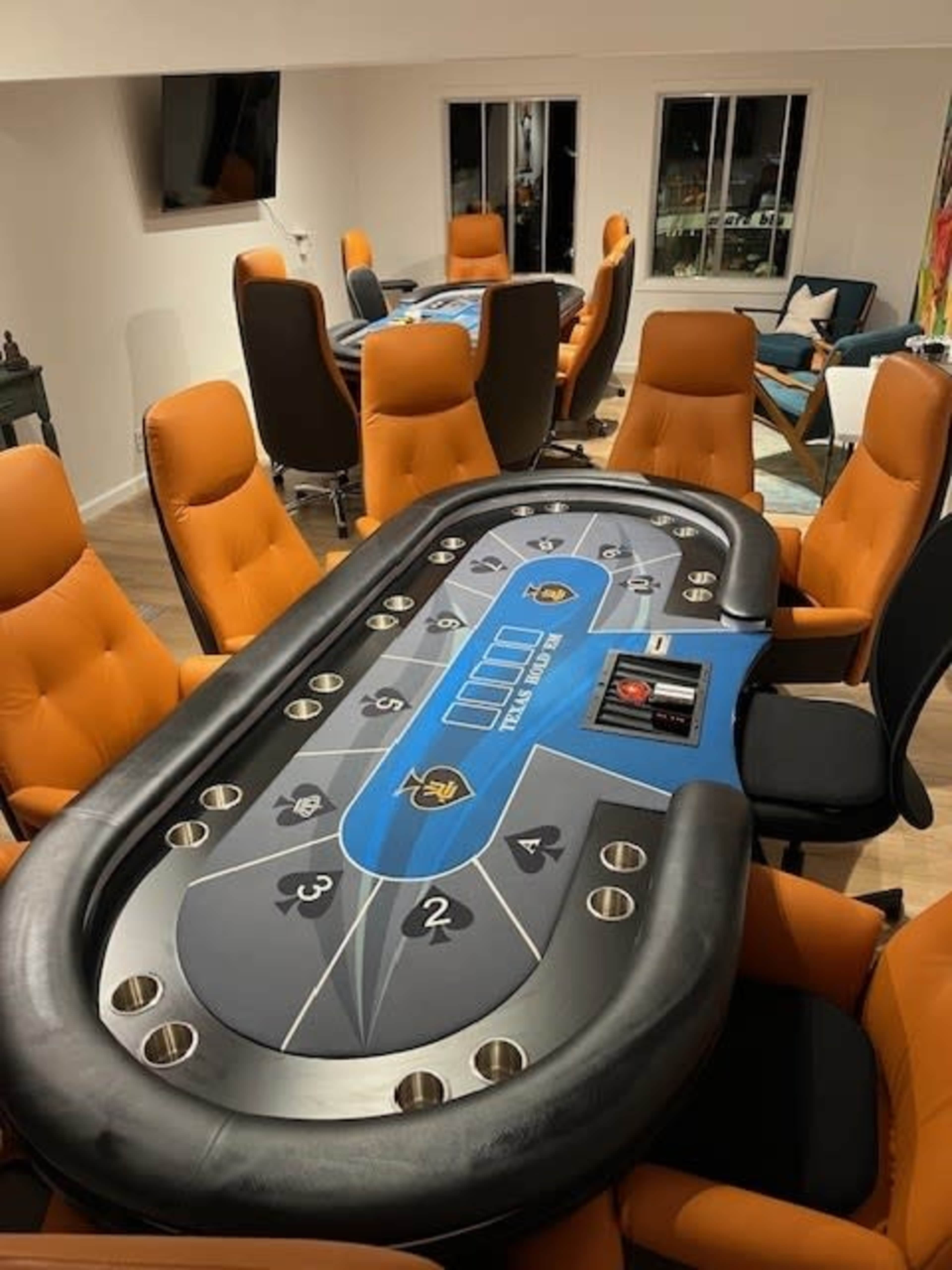 A poker table with a blue and black layout is surrounded by orange chairs in a well-lit room.