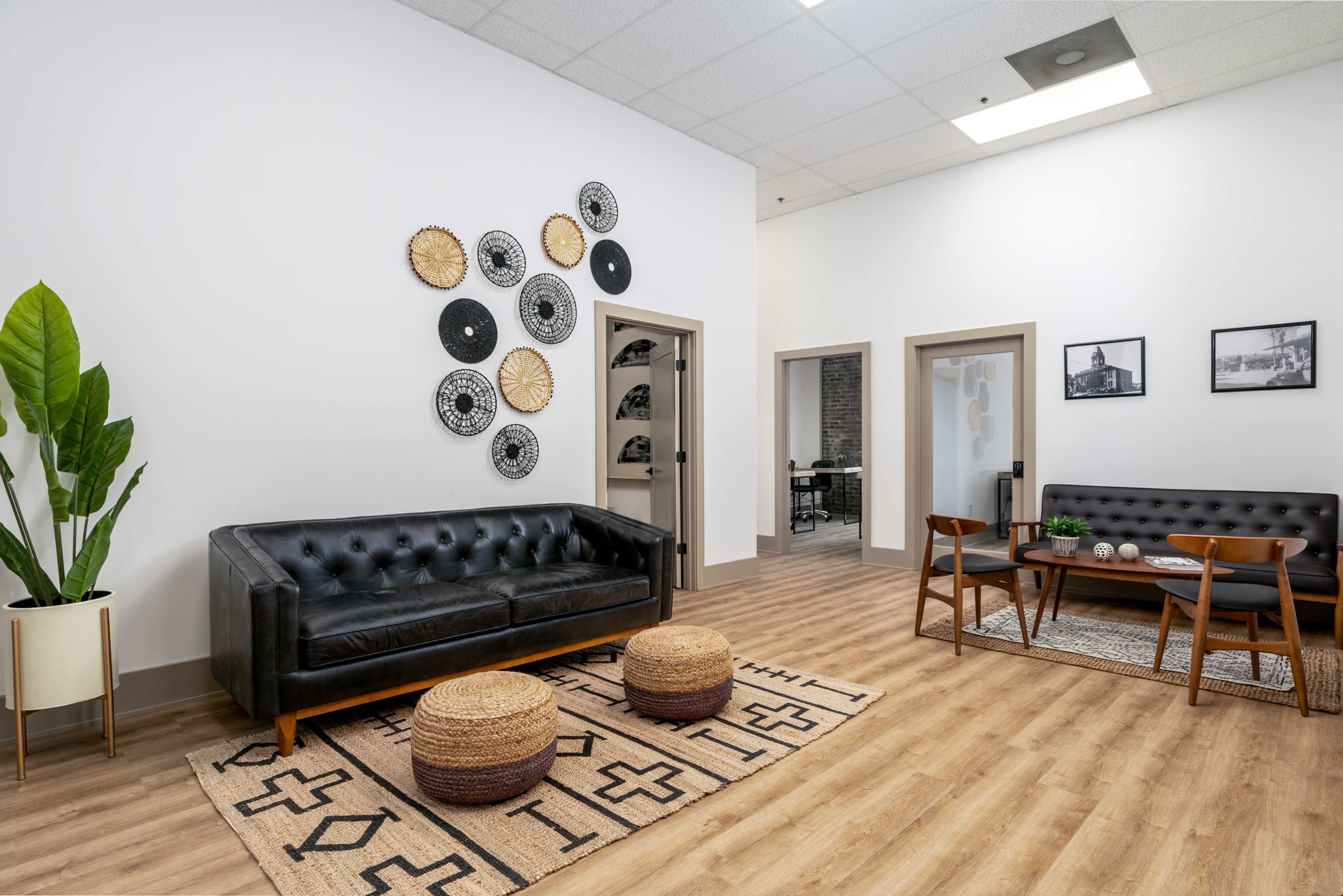 A modern waiting area features a black tufted sofa, a patterned area rug, two round poufs, and wall decor arranged above a small dining table set.
