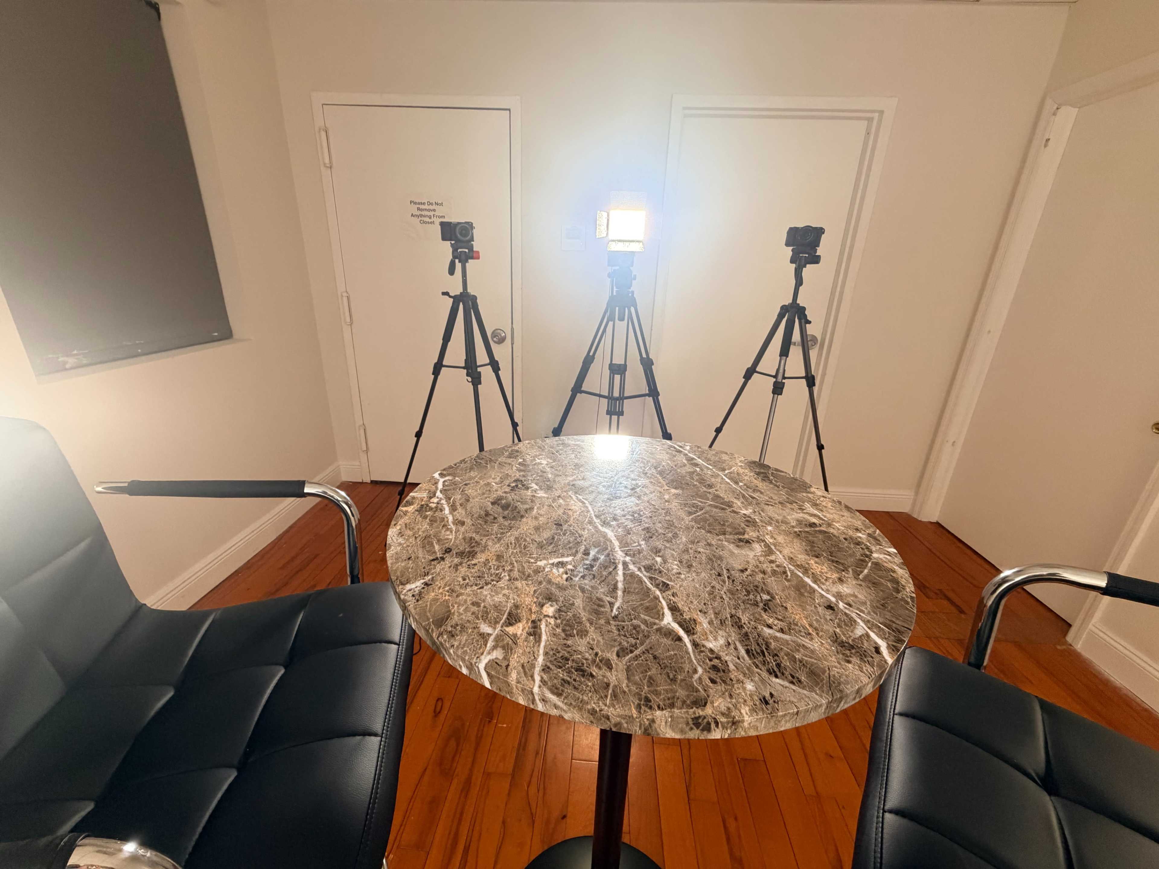The image shows a small room with a round marble table and two black chairs, flanked by three camera tripods, two with lights, facing the table.