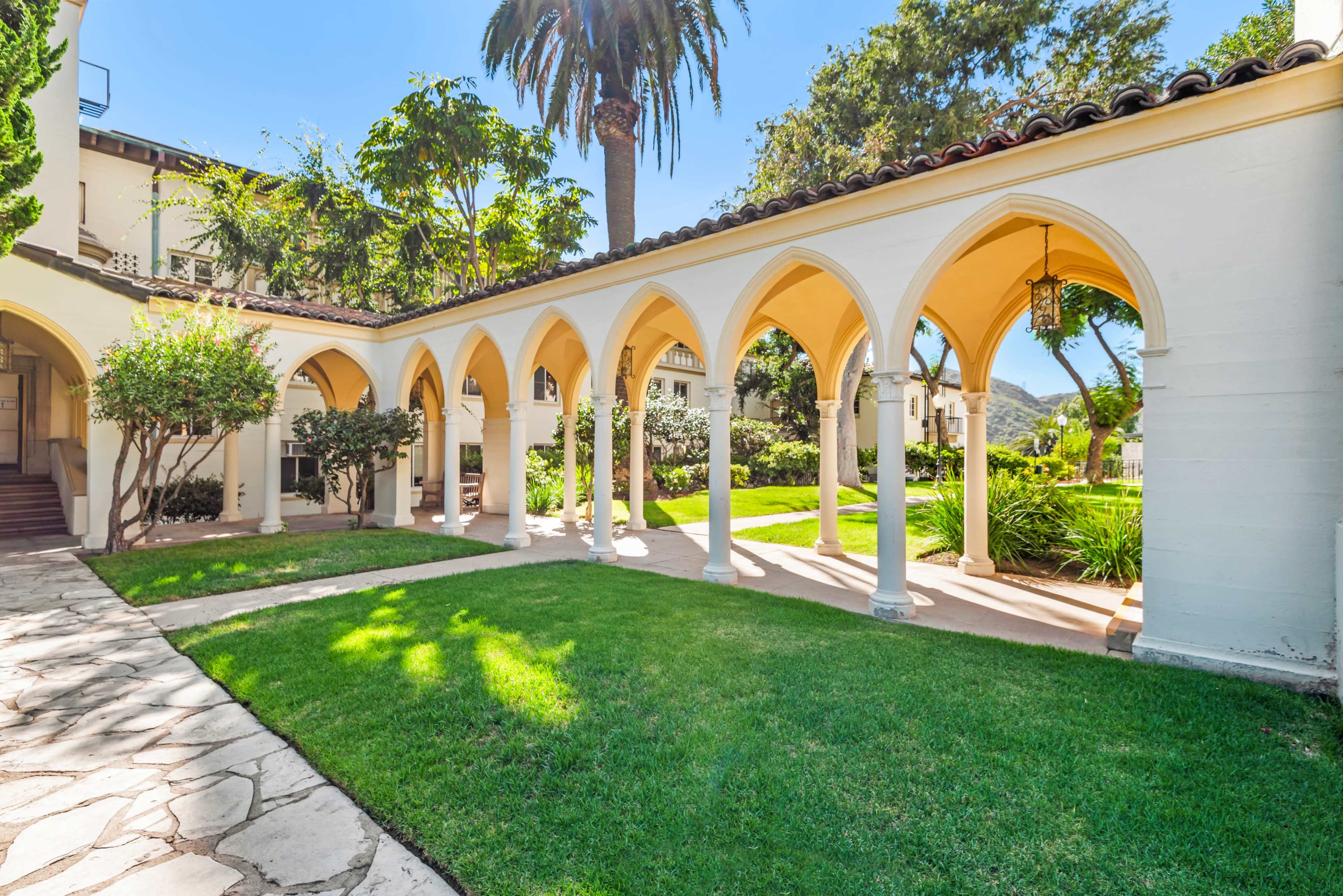 The image shows a colonnade with arches leading to a landscaped garden surrounded by greenery and palm trees.