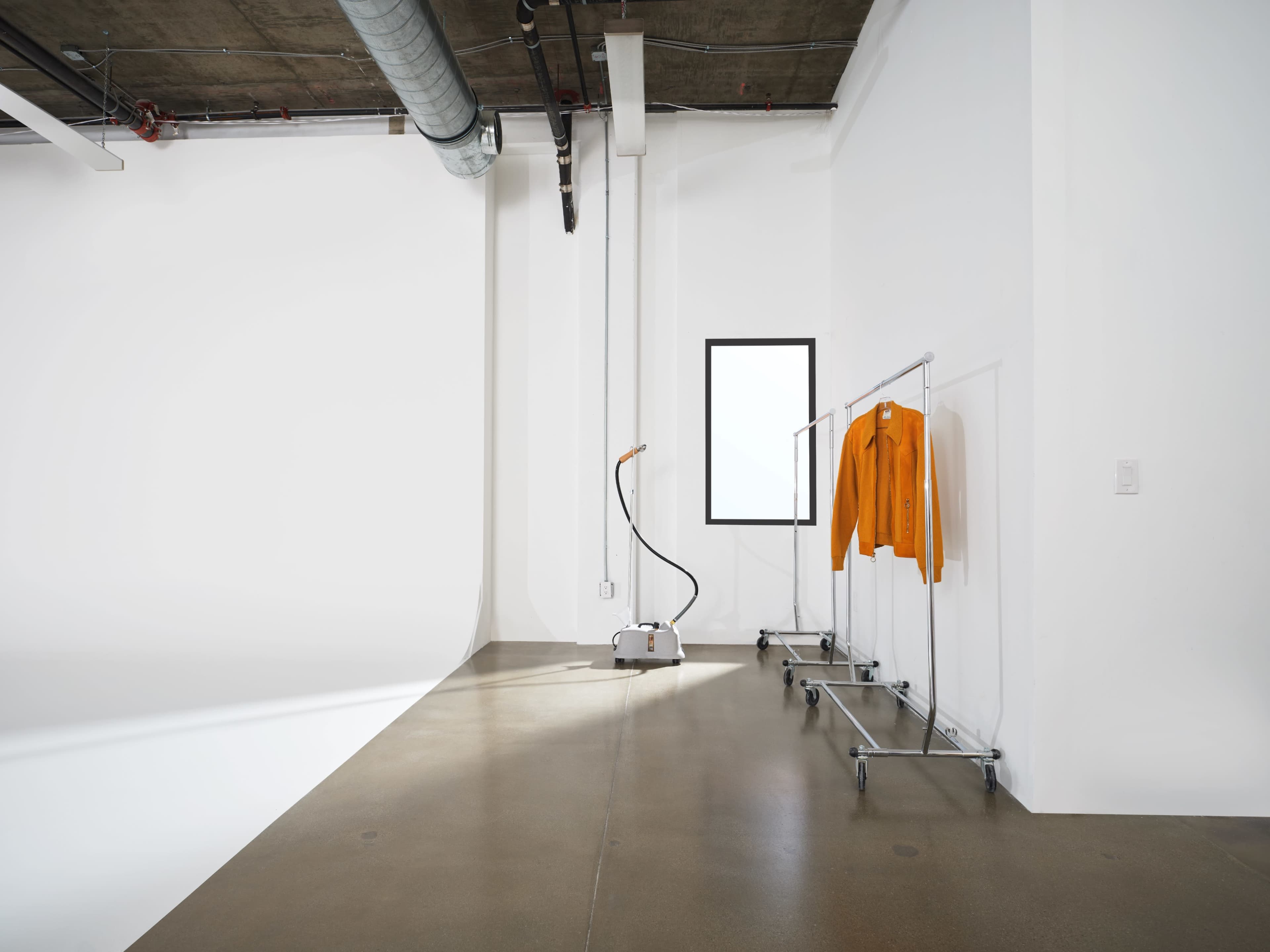 A minimalist studio space with a window, a garment steamer on the floor, and an orange jacket hanging on a clothing rack.