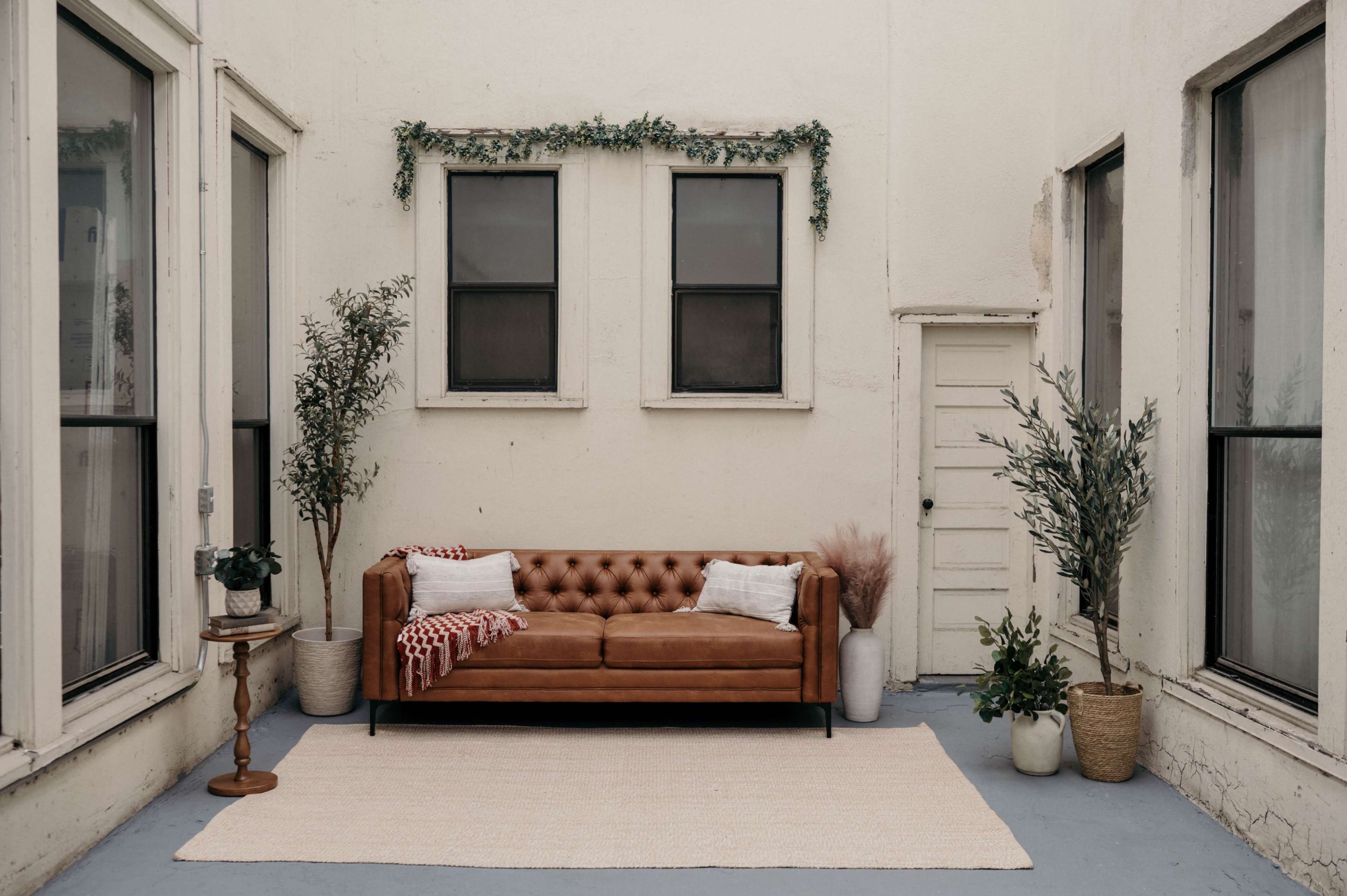 The image shows a cozy indoor space featuring a brown leather couch, two potted plants, and a small table, all set against white walls and large windows.