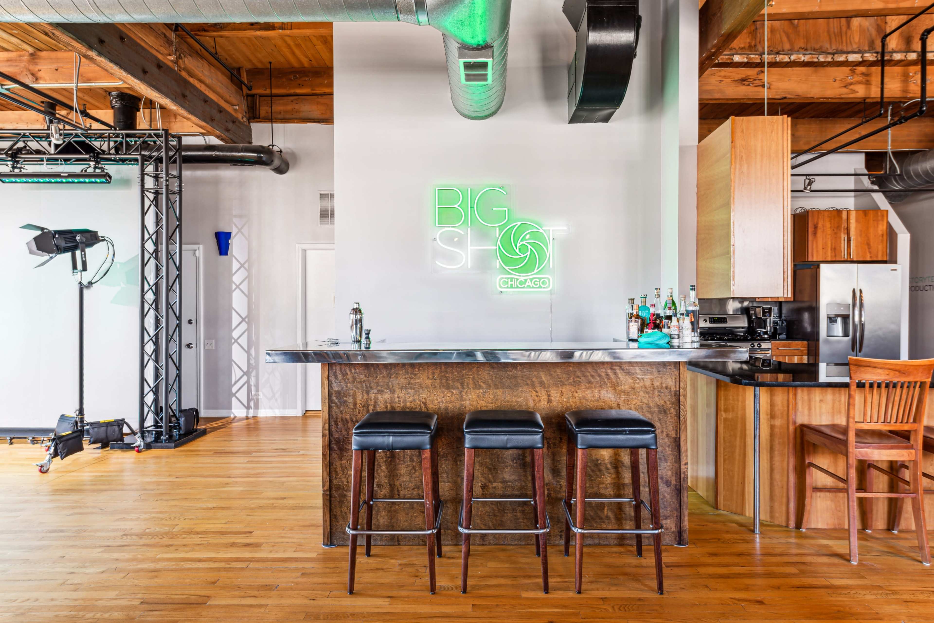 A modern bar area with a wooden countertop and black stools, featuring a neon sign that reads "BIG SPOT CHICAGO" on the wall.