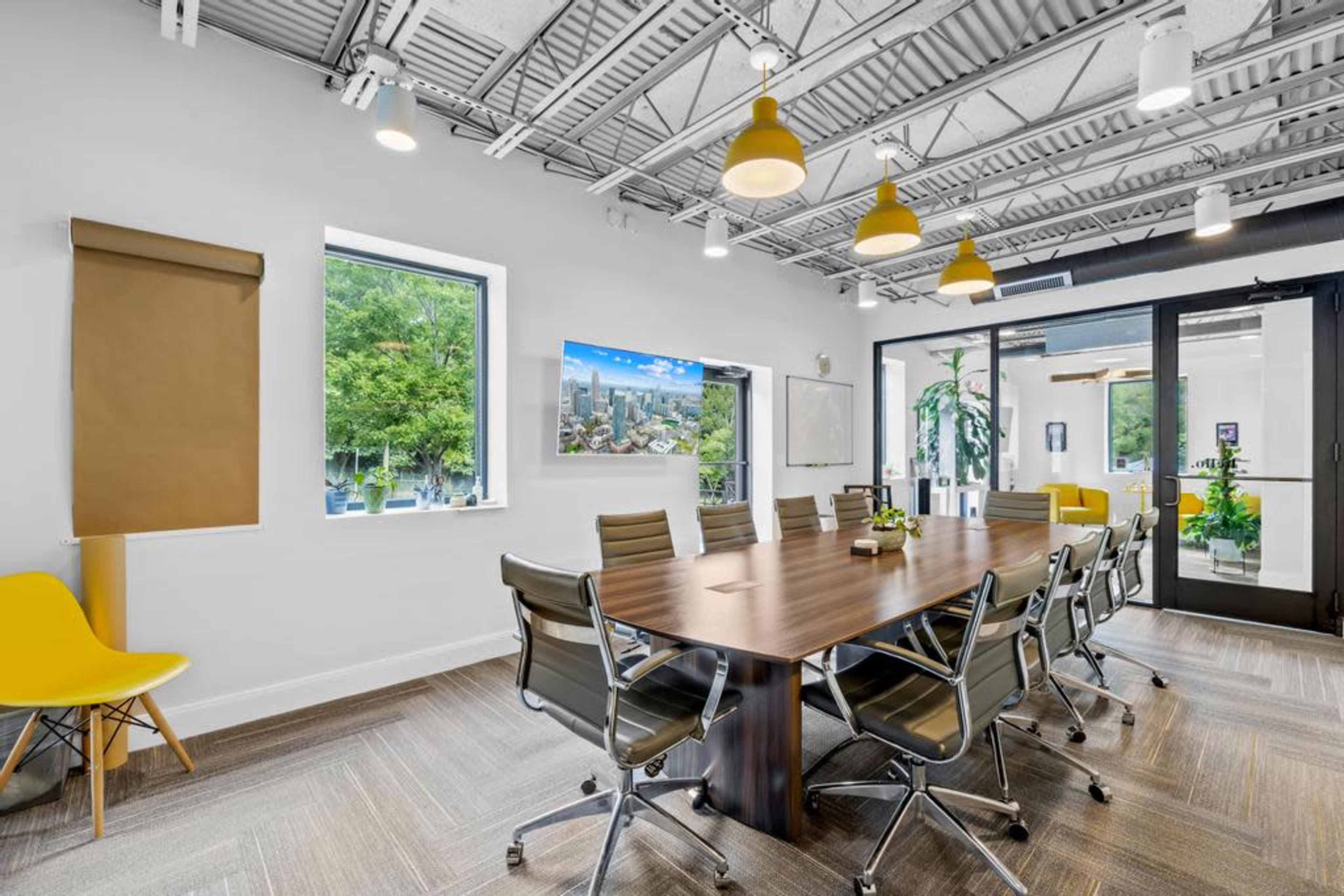 A modern conference room features a large wooden table surrounded by ergonomic chairs, with windows overlooking greenery and a television displaying a cityscape.