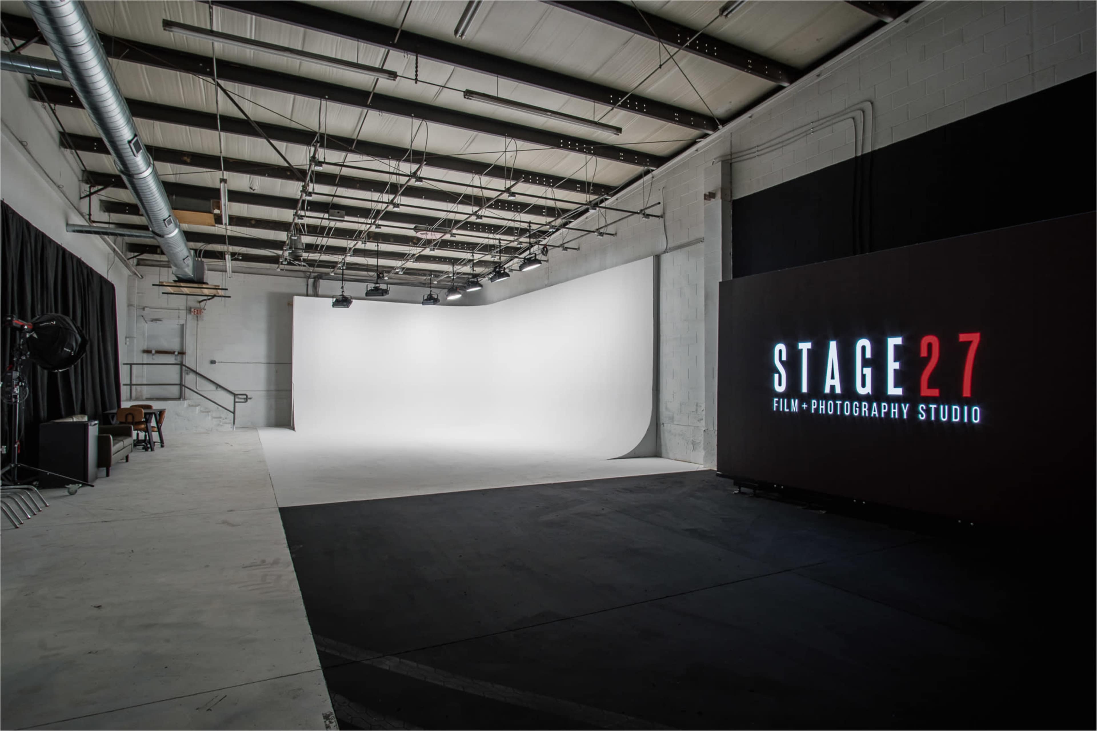 An empty photography studio featuring a white backdrop, black flooring, and a large illuminated sign reading "STAGE27 FILM + PHOTOGRAPHY STUDIO."
