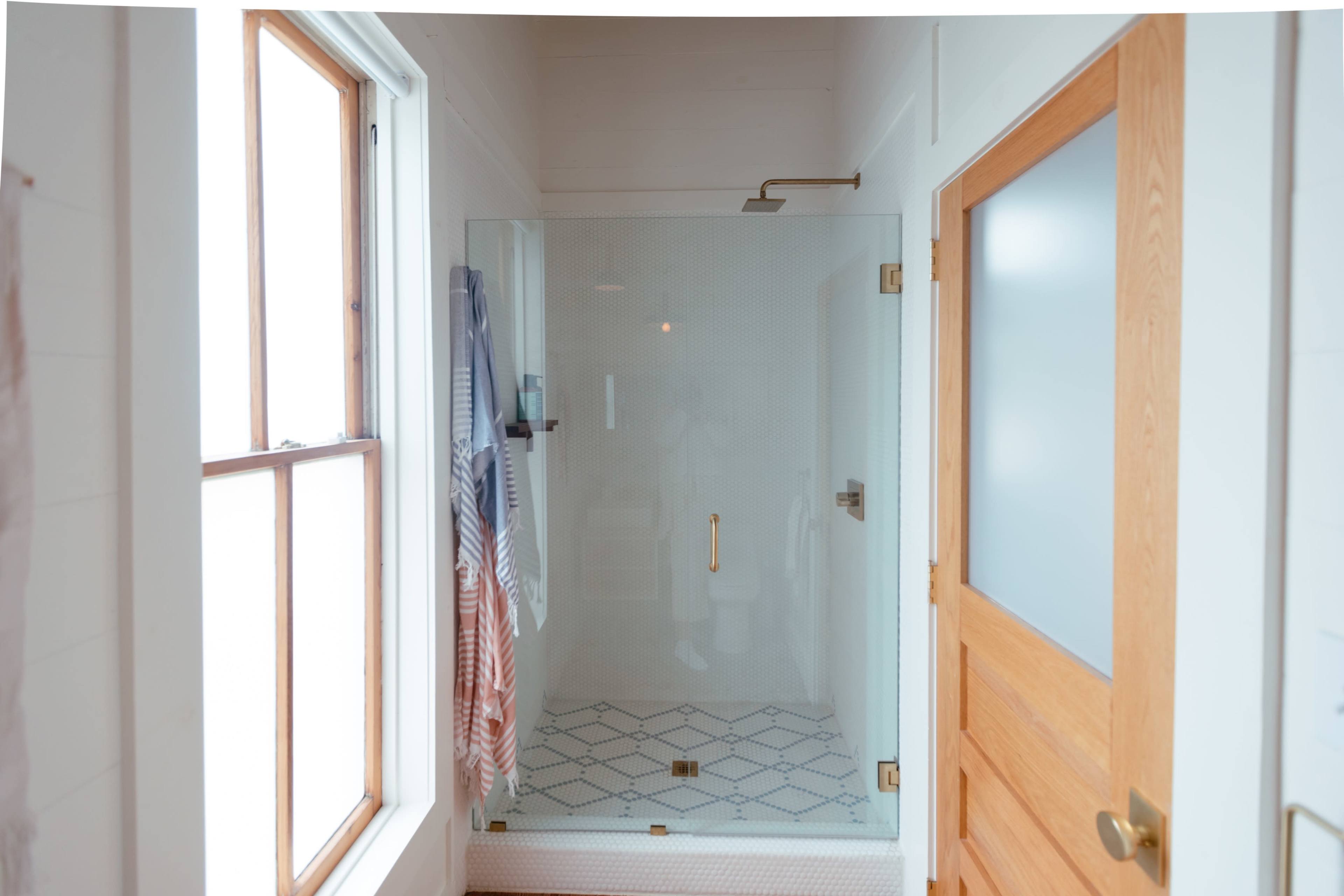 A clear glass shower enclosure stands in a bathroom next to a wooden door and windows, with towels hanging nearby.