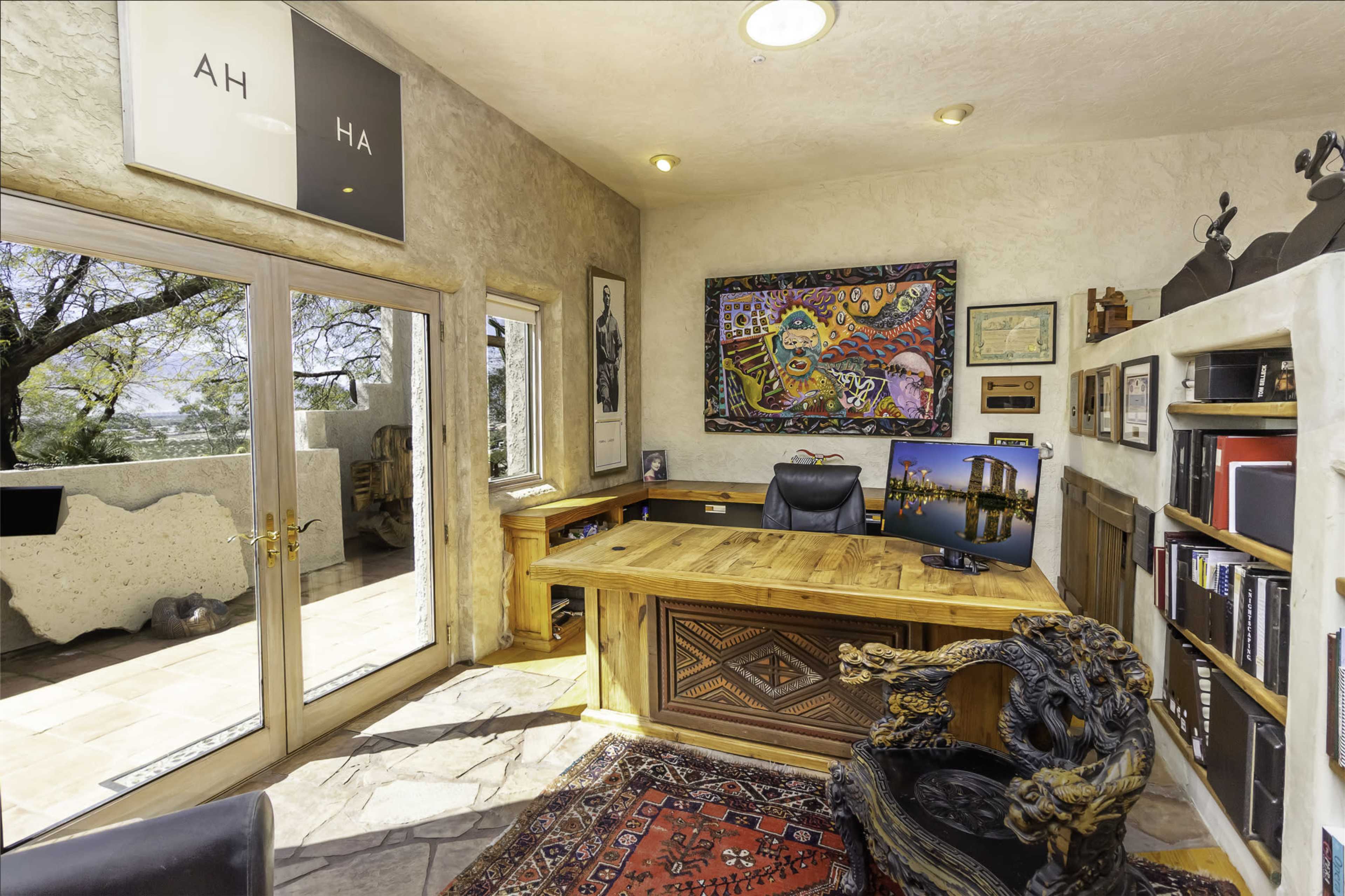 A well-furnished office with a wooden desk, a colorful artwork on the wall, and large windows overlooking a patio.