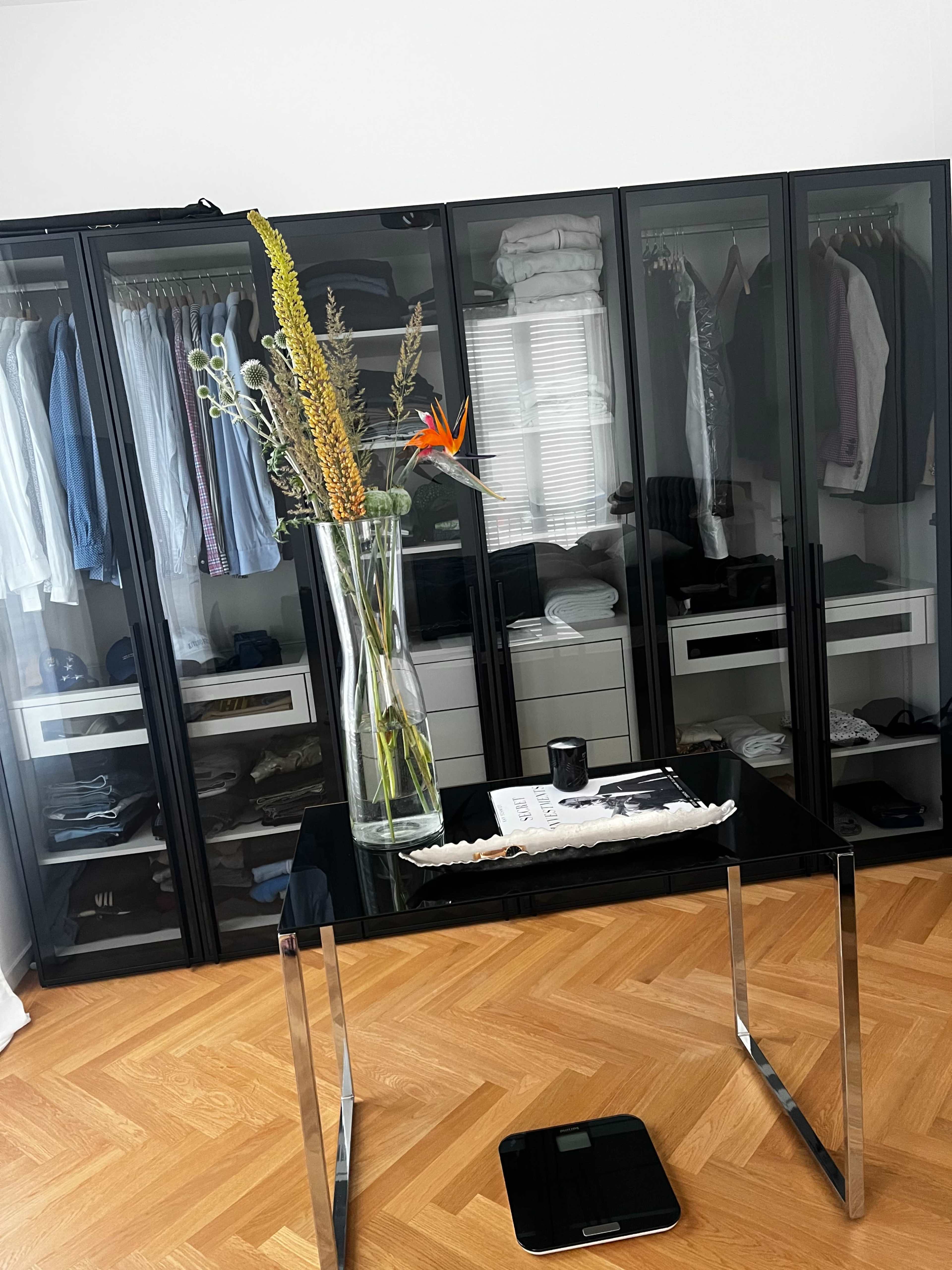 A modern room with a glass table and a tall vase of flowers in front of a wardrobe featuring neatly organized clothes.