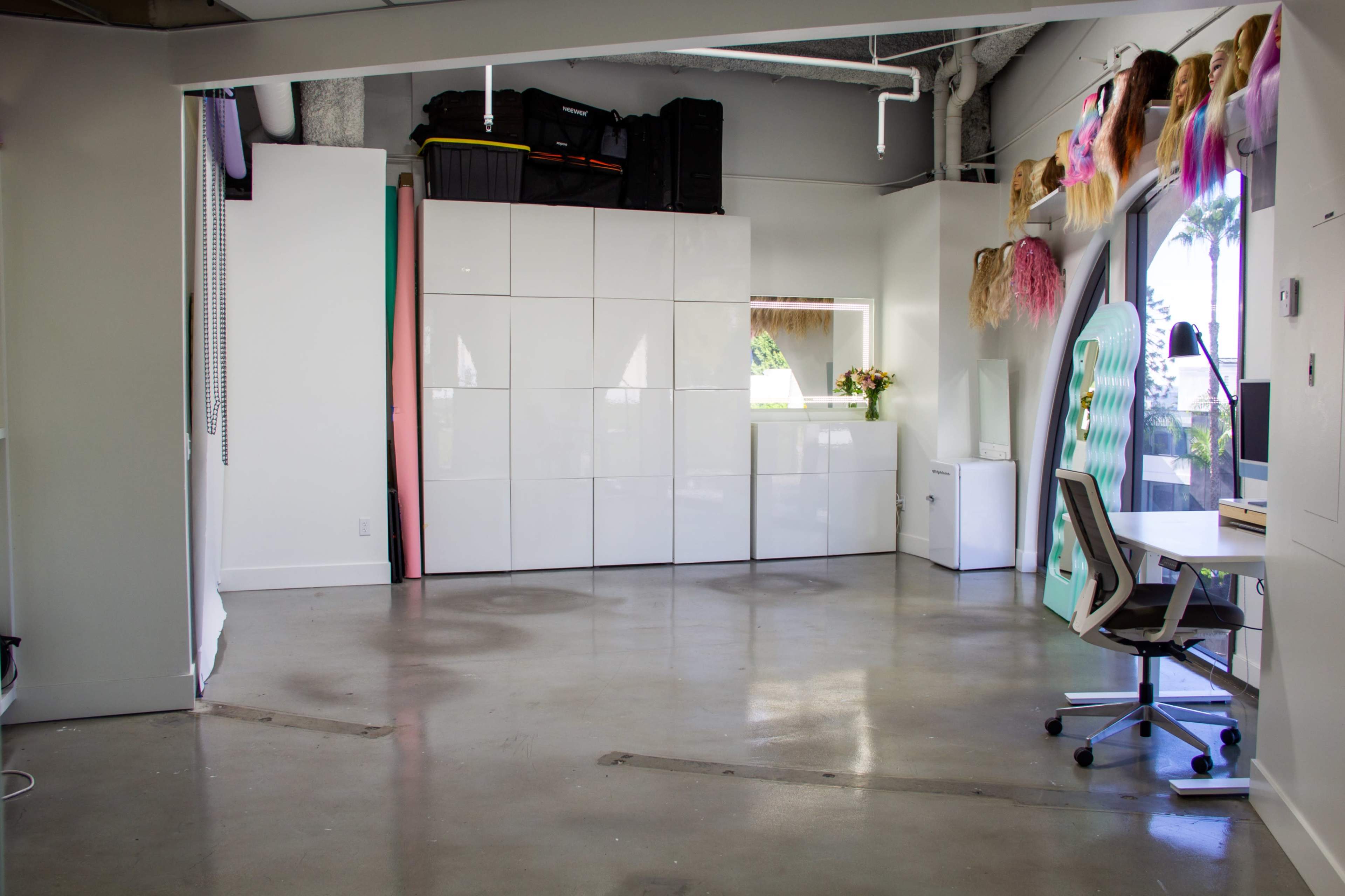 The image shows a spacious room with white walls, polished concrete floors, and a large white storage unit, featuring a desk with a chair and decorations of colorful hairpieces on the wall.