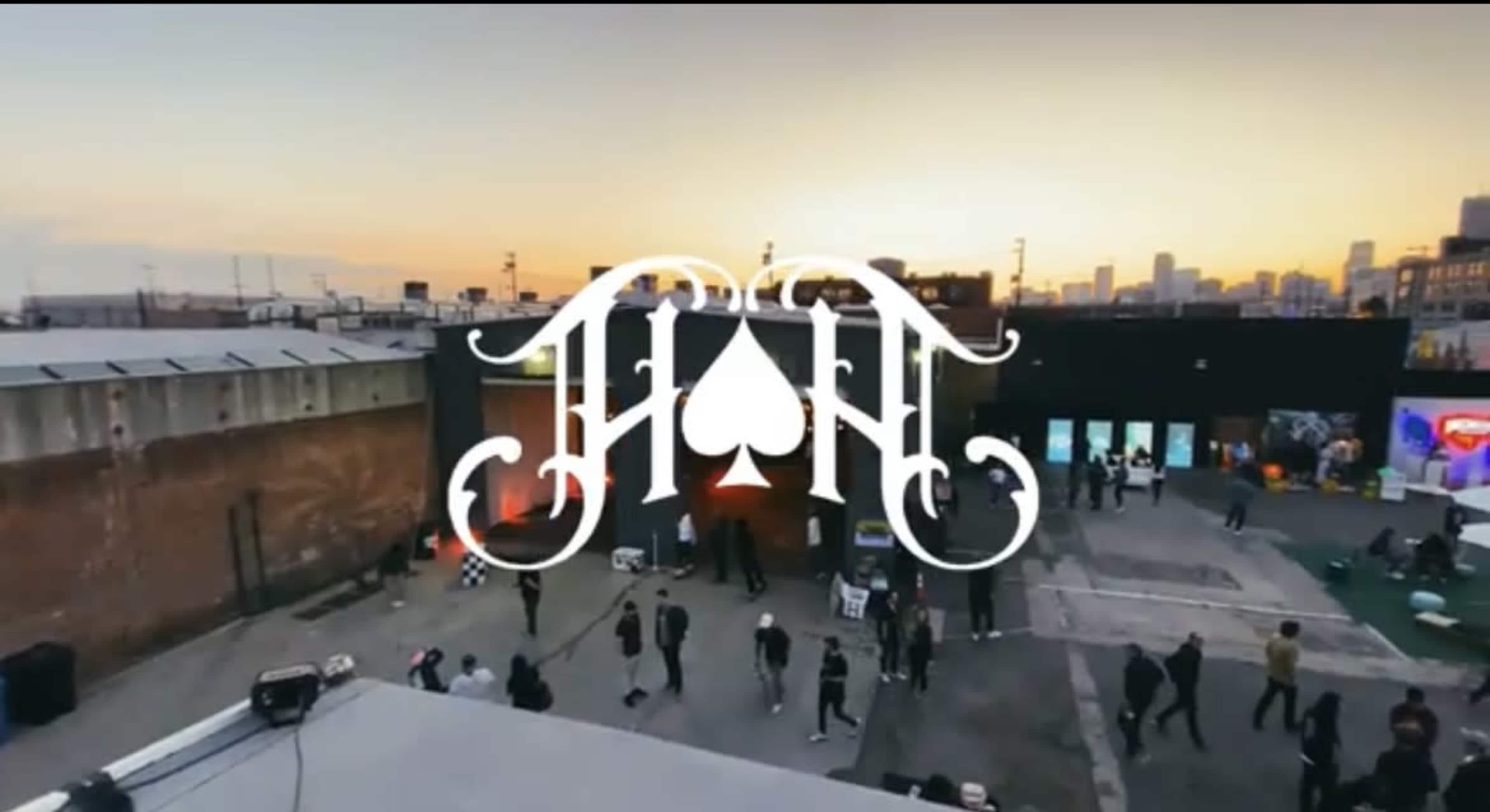 A crowd gathers outside a venue with a skyline view at sunset, featuring a prominent logo displaying a heart and decorative elements.