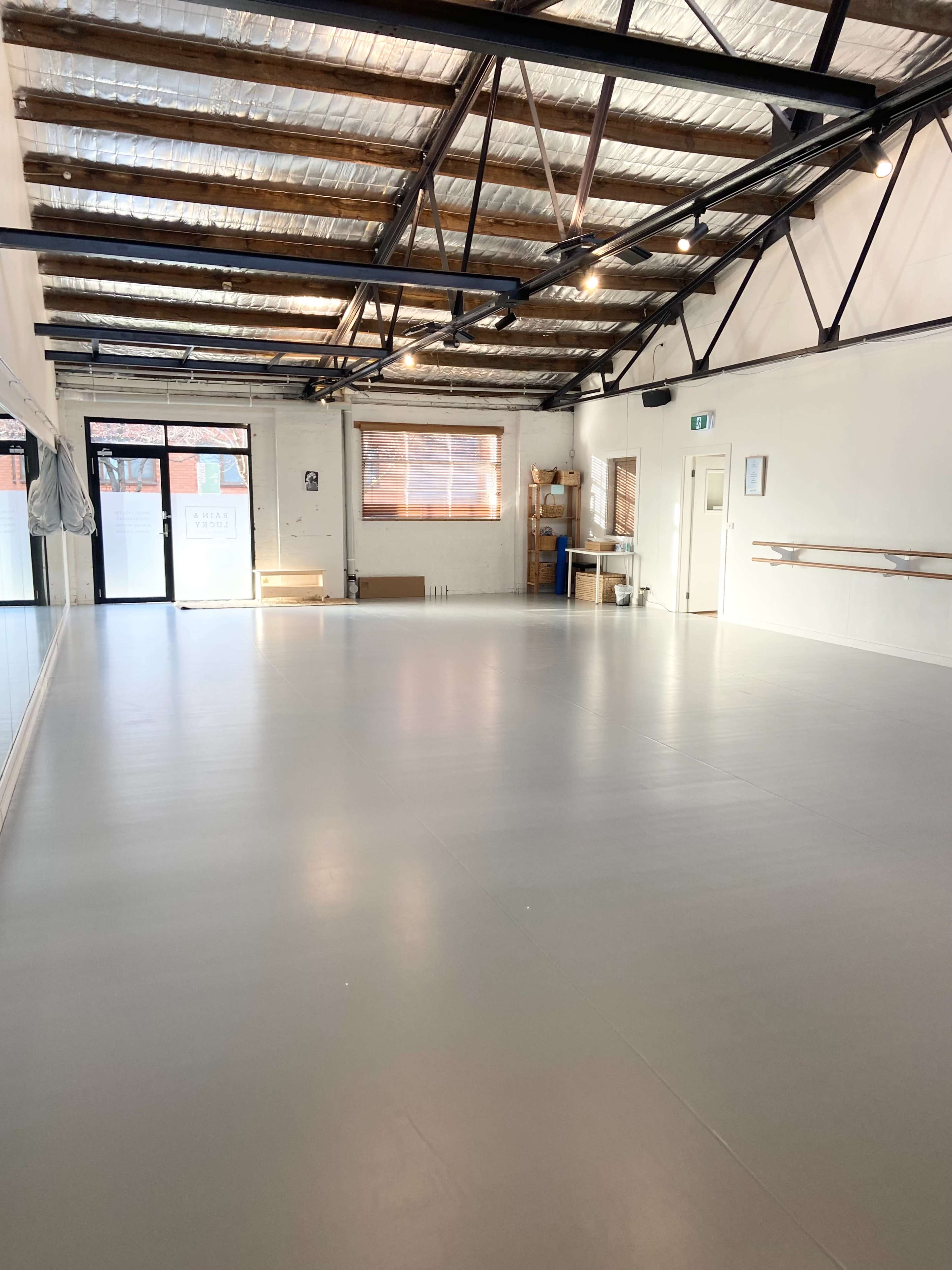 The image shows a spacious, empty dance studio with gray flooring and large windows letting in natural light.