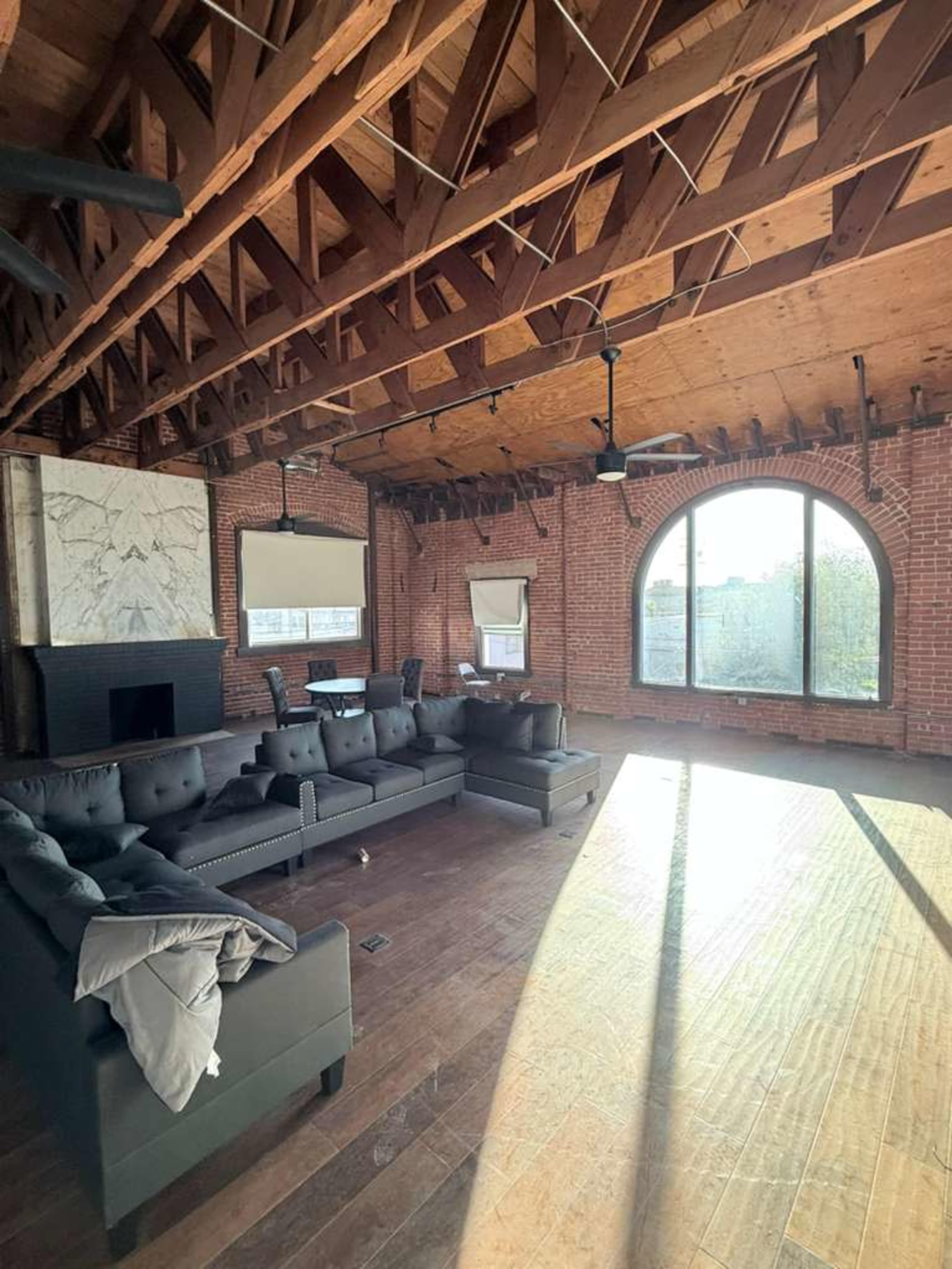 The spacious living area features a large sectional sofa, exposed brick walls, high wooden ceilings, and a prominent arched window.