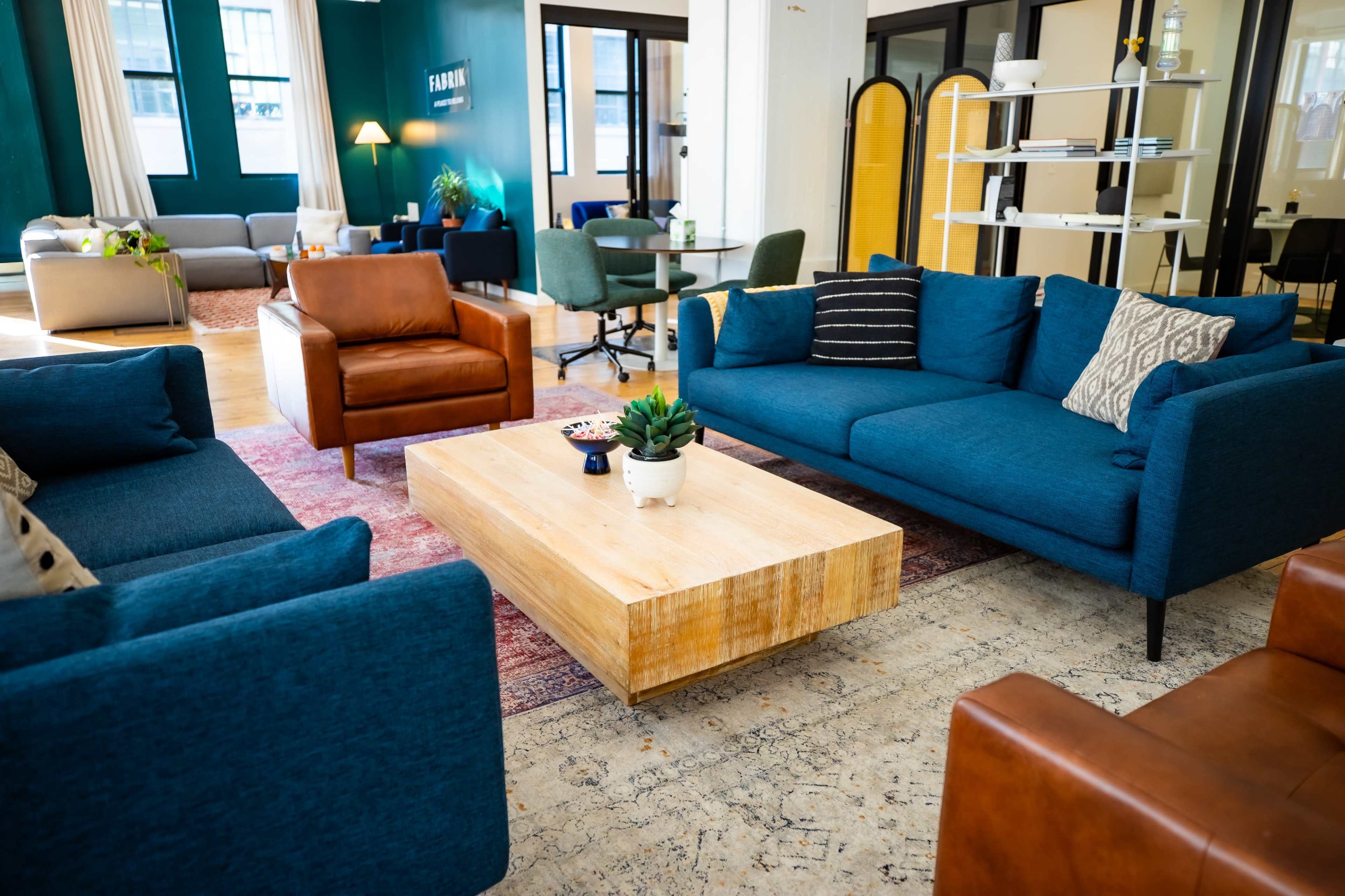 The image shows a modern lounge area with blue couches, a wooden coffee table, and a mix of seating arrangements.