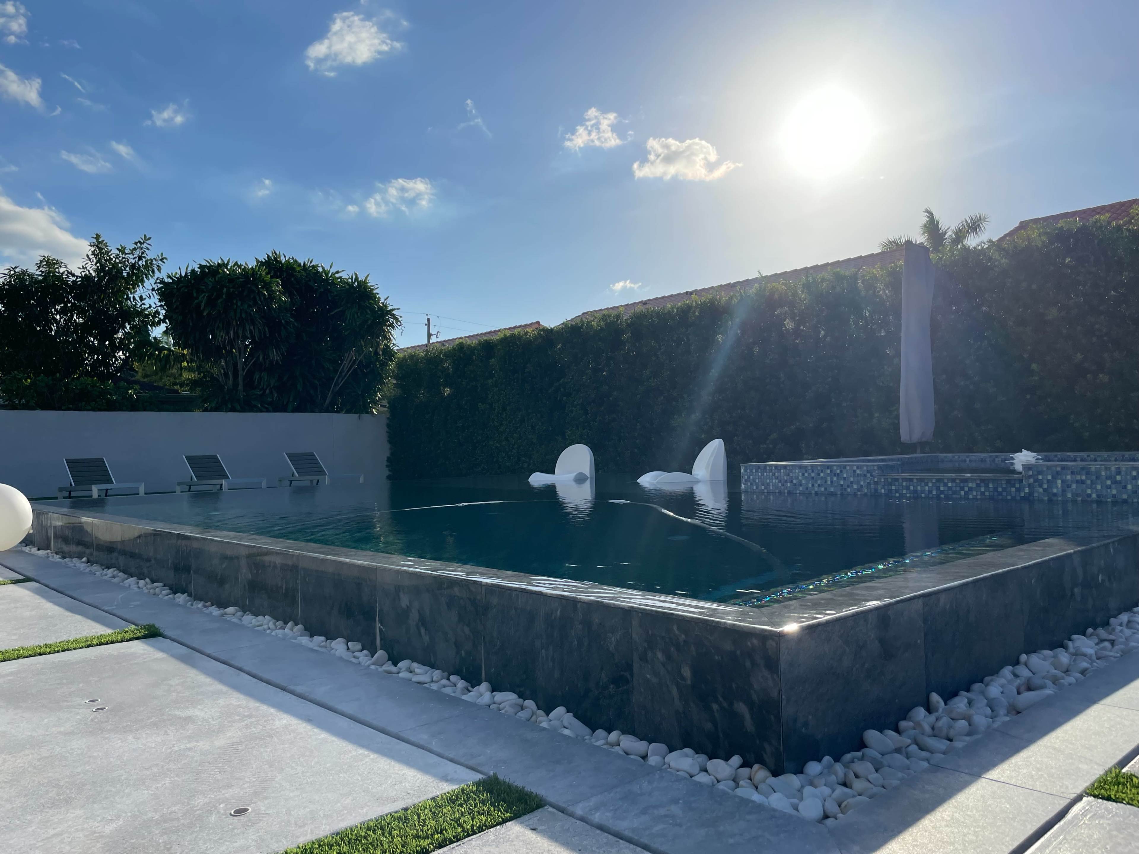 The image shows a modern swimming pool surrounded by concrete flooring, lounge chairs, and landscaped greenery under a clear blue sky.