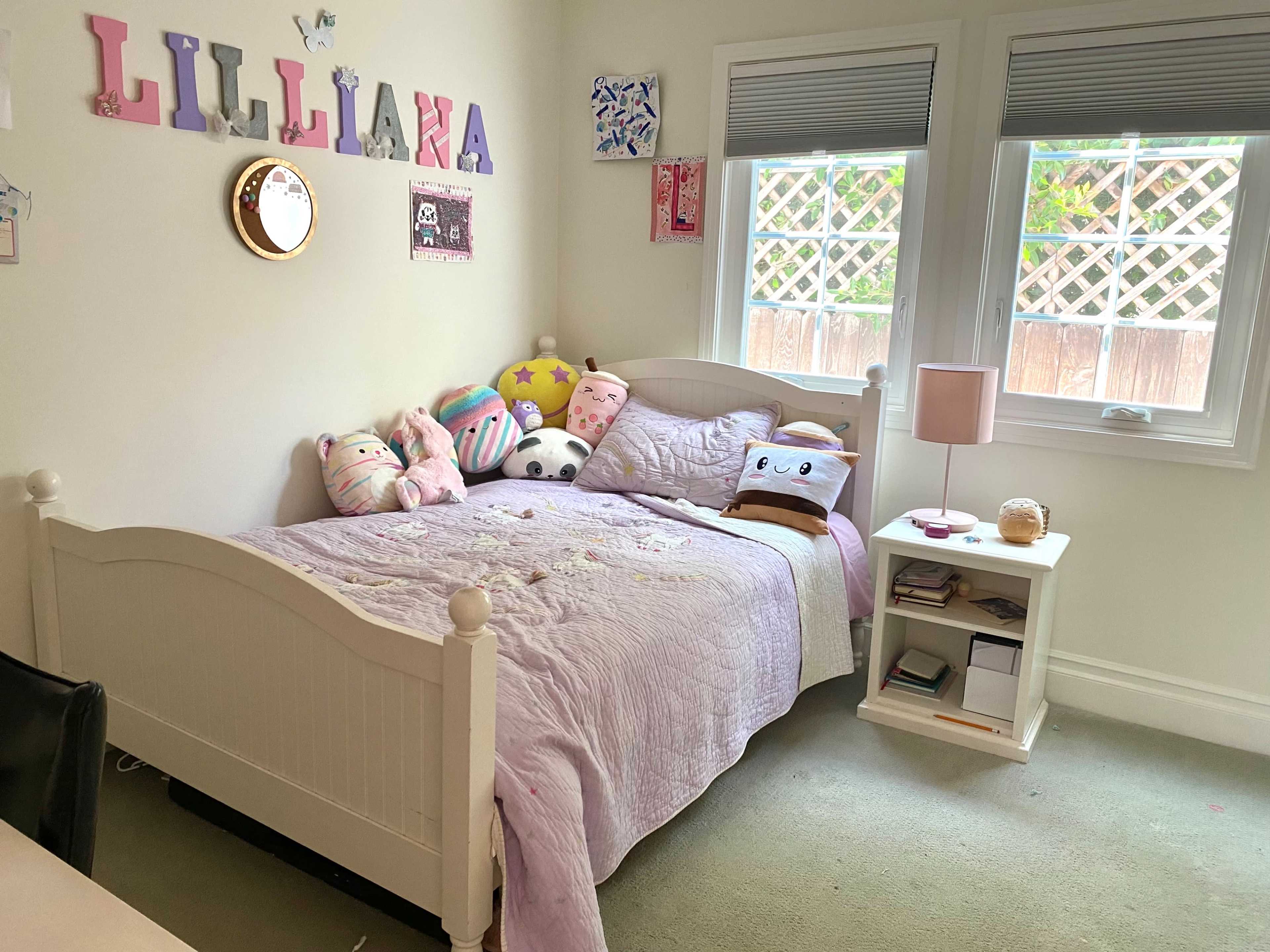 A bedroom with a bed, decorated with plush pillows, a nightstand, and a wall adorned with the name "Lillian" made of colorful letters.