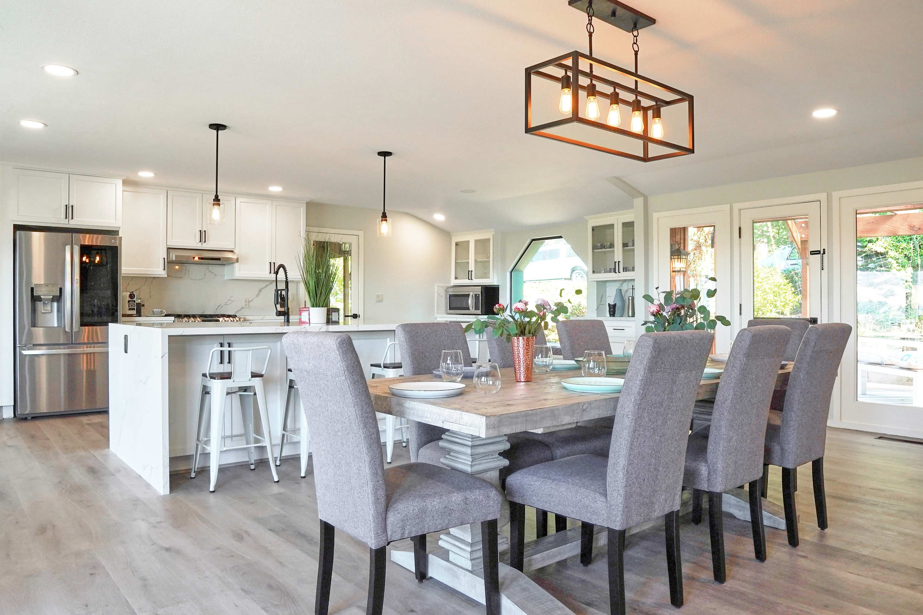 The image shows a modern kitchen and dining area featuring a large wooden table with gray upholstered chairs and pendant lighting above.