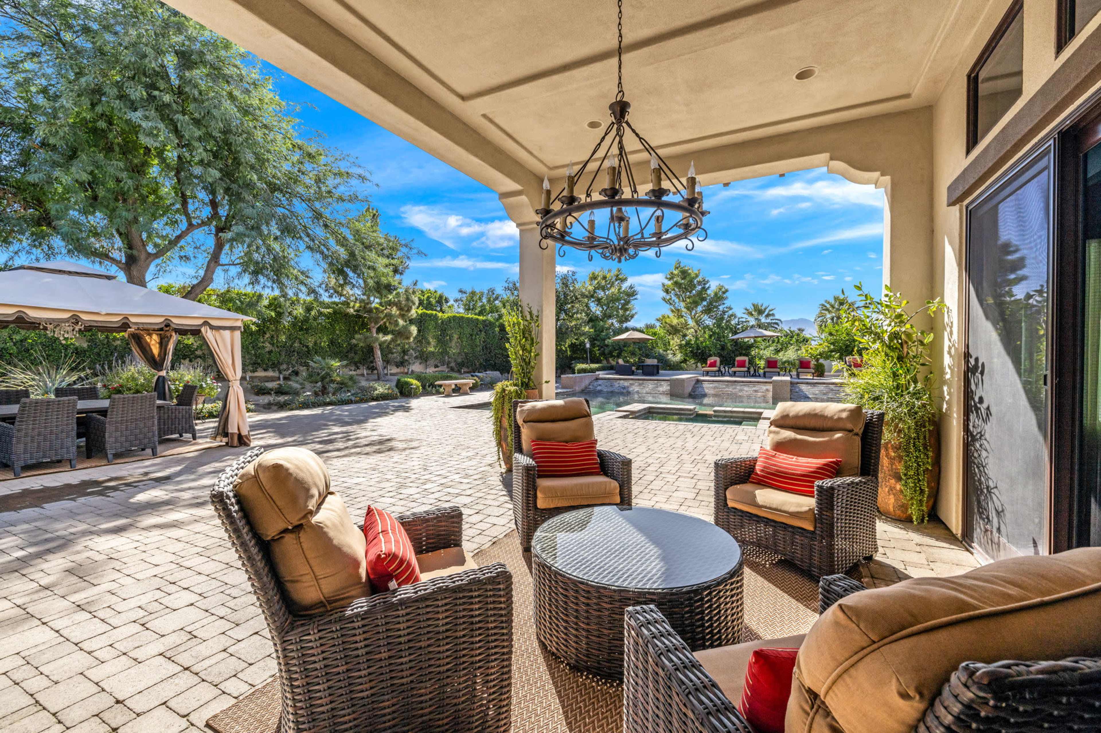 A spacious patio area with wicker chairs around a round table, surrounded by a landscaped garden and a swimming pool in the background.