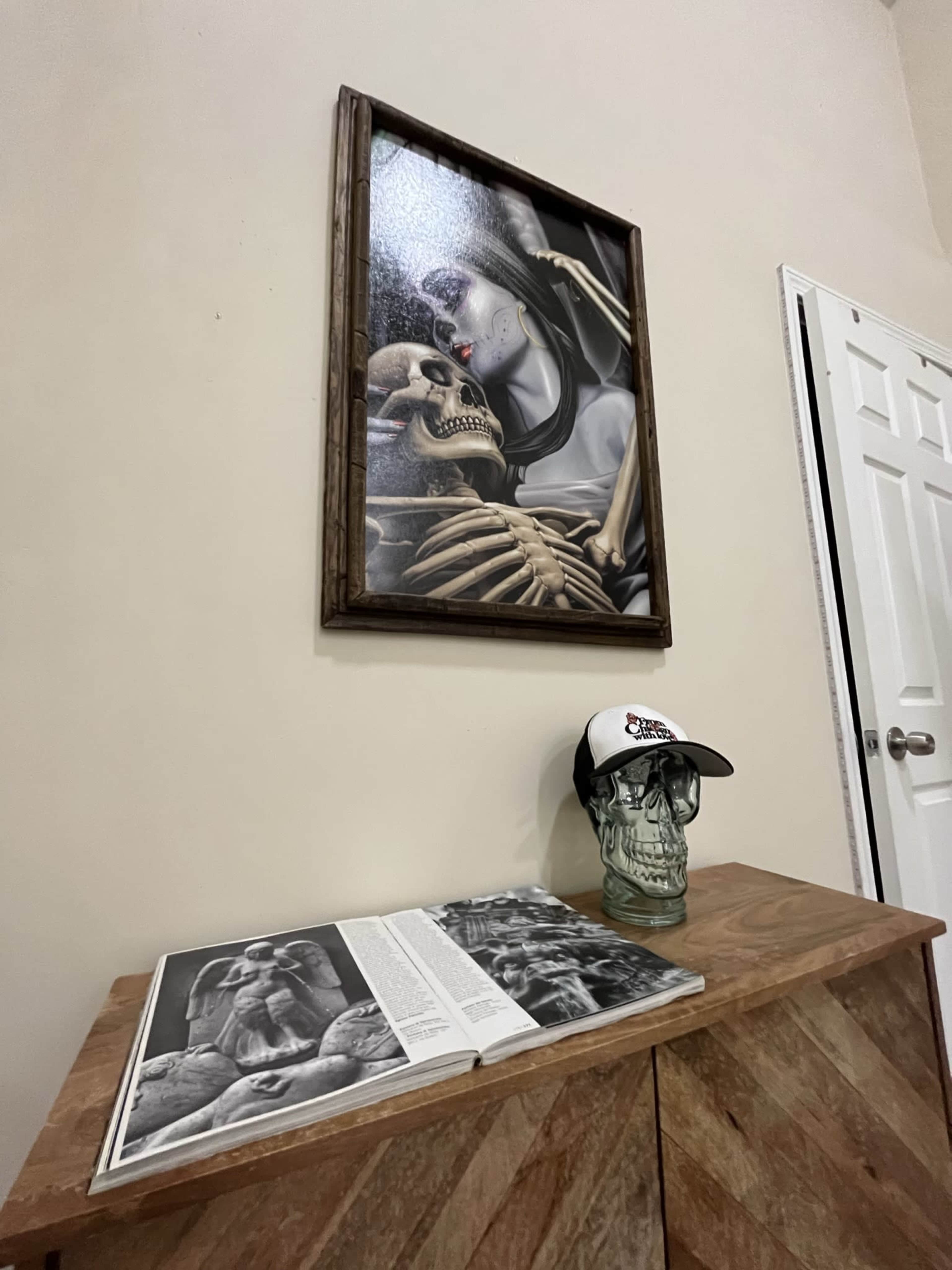 A wall with a framed painting of a skeleton and a figure, above a wooden dresser displaying an open book and a skull-shaped object wearing a cap.