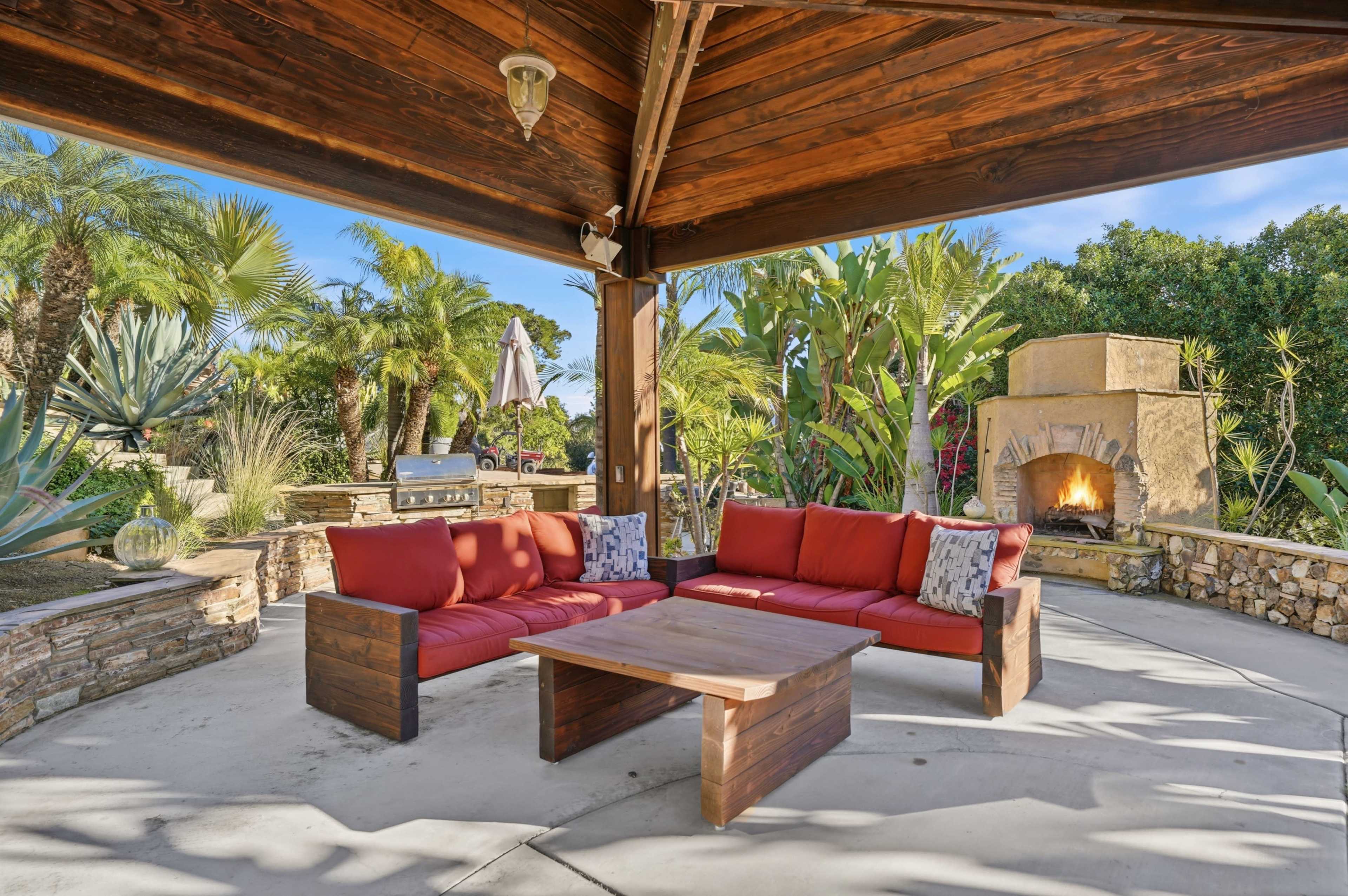 The image shows a covered outdoor seating area featuring a rectangular wooden table and red cushioned sofas, surrounded by various plants and a stone fireplace.