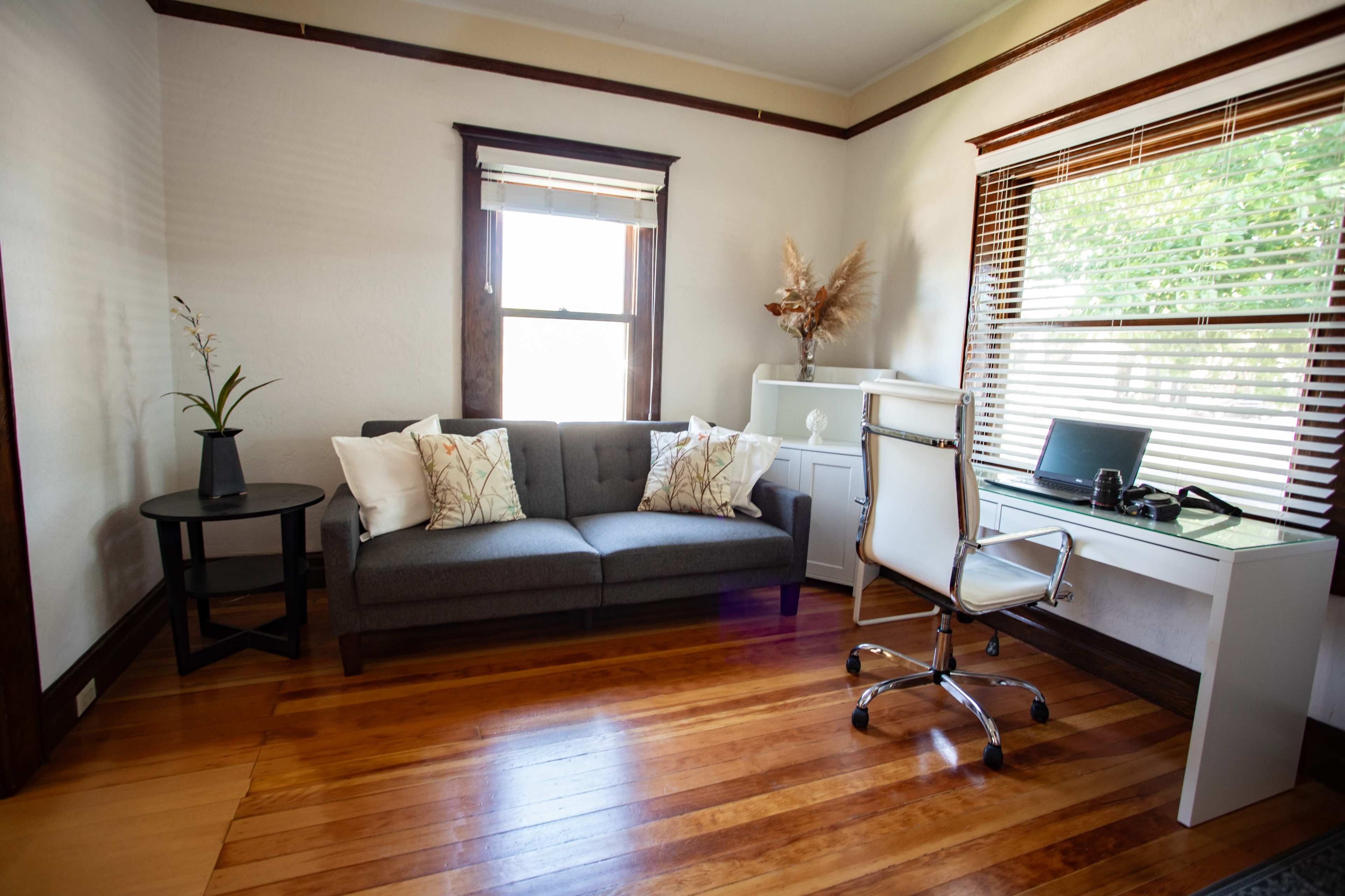 The image shows a modern office space with a gray couch, a round black side table, a white desk with a computer, and large windows allowing natural light to enter.