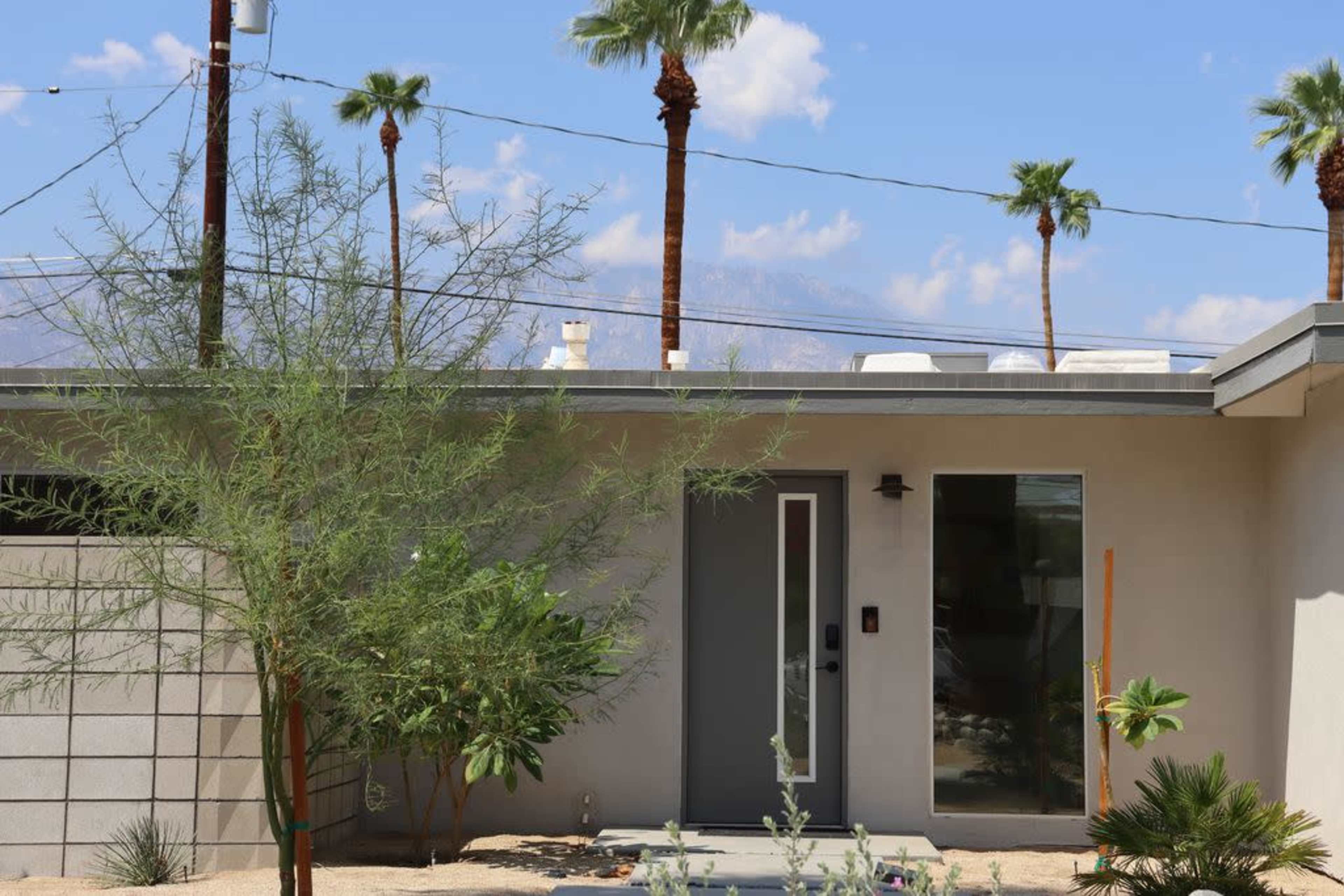 A modern house with a flat roof and large front door is surrounded by desert plants and palm trees against a backdrop of mountains and blue sky.