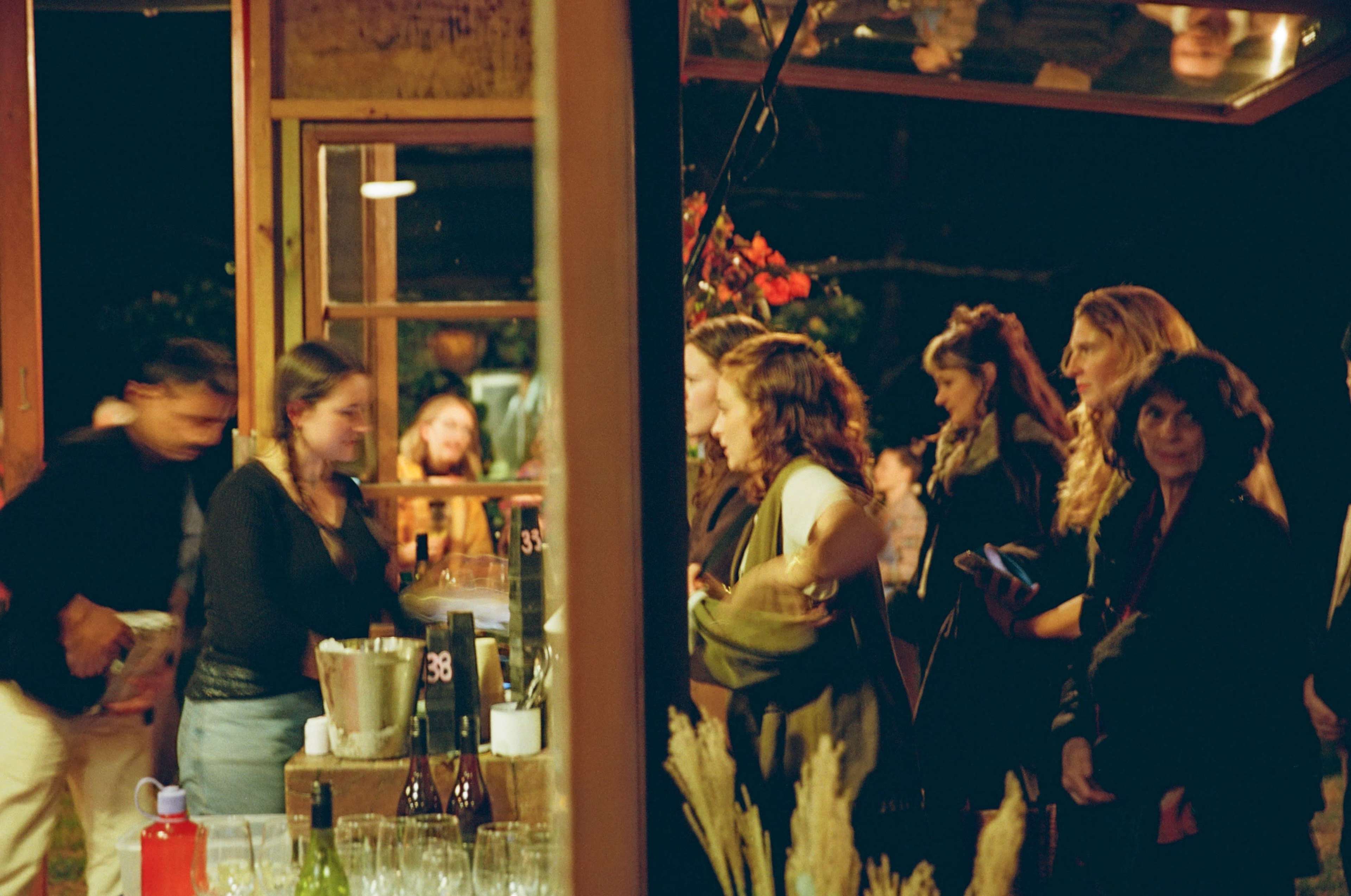 A group of people socializes at a bar during a nighttime gathering, with some standing near the counter and others in conversation.