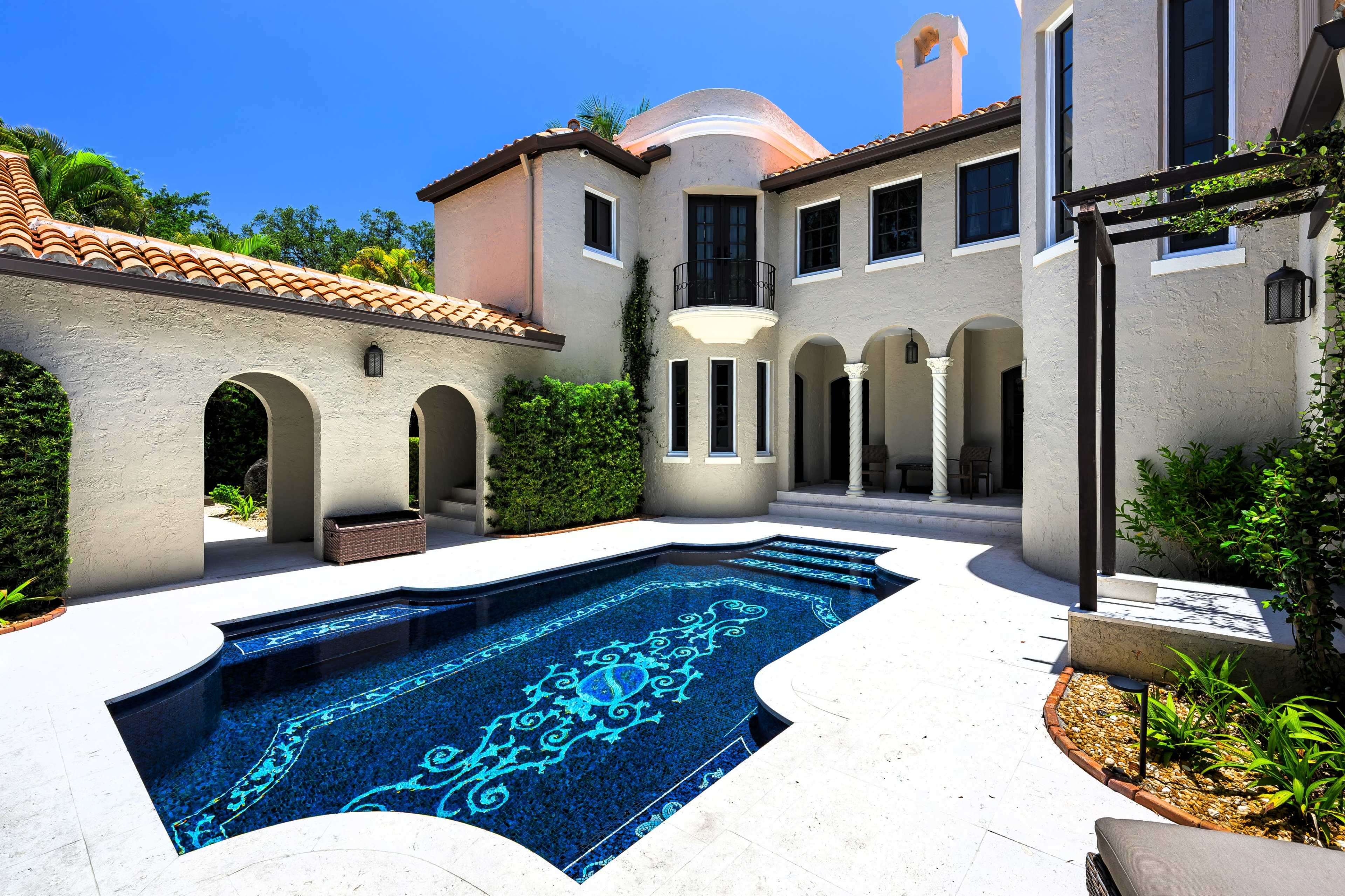 The image depicts a luxury home with a tiled swimming pool surrounded by lush greenery and architectural features, including arched doorways and a tiled roof.