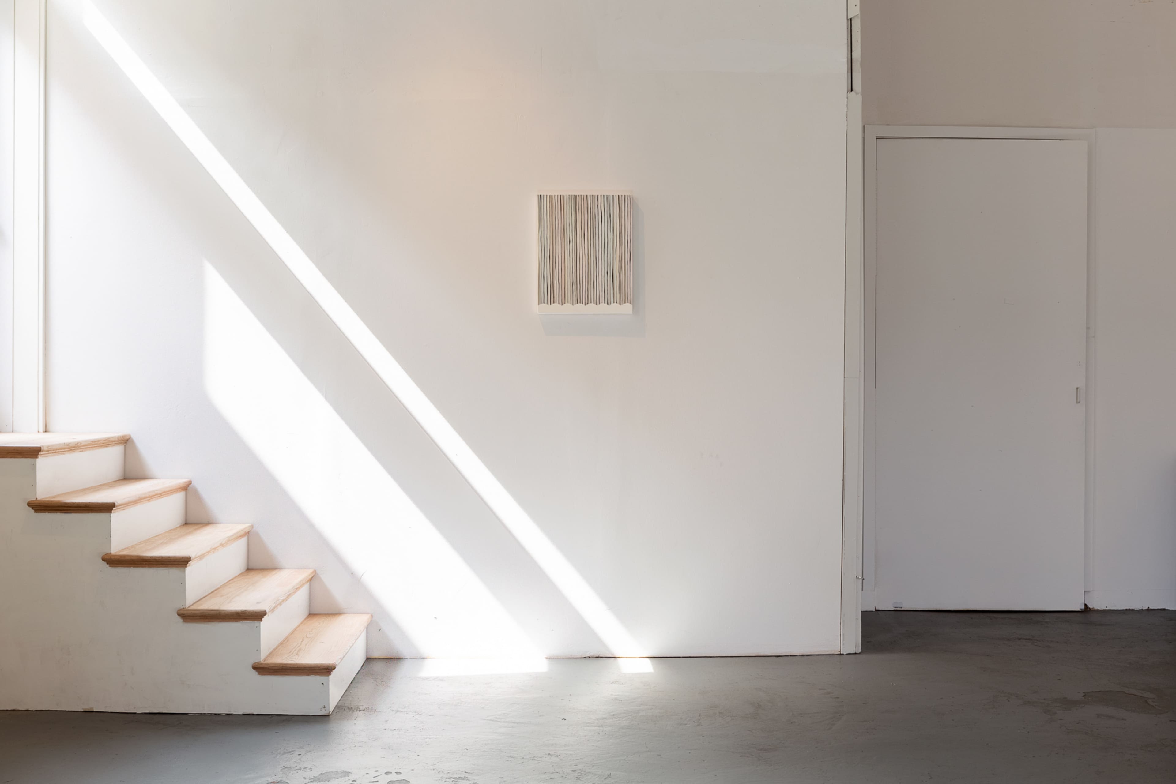 A staircase leads up to a white wall with a framed piece of art, illuminated by a diagonal beam of light.