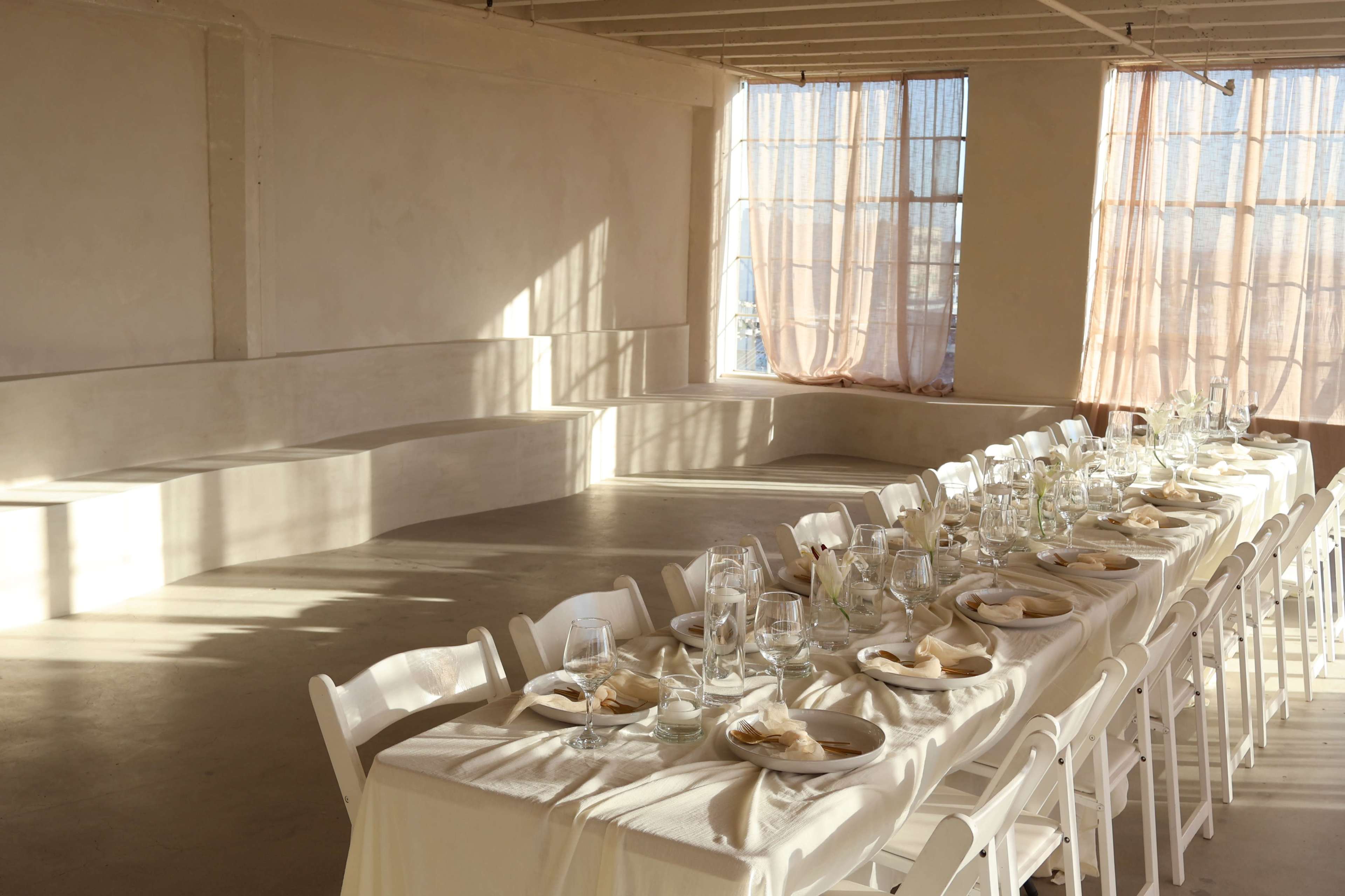 A long dining table is set with glassware and plates in a bright, minimalistic room with large windows and soft curtains.