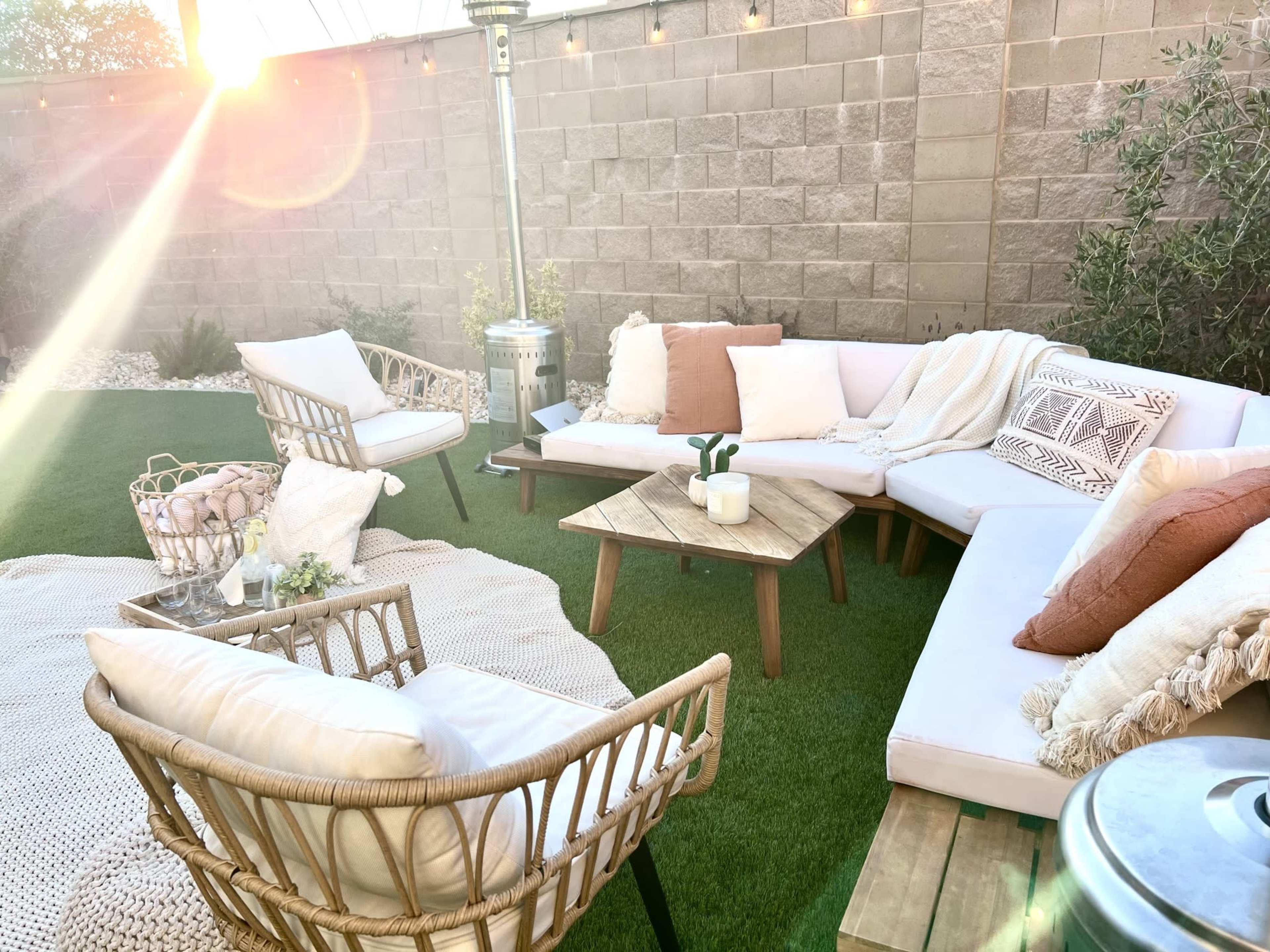 A cozy outdoor seating area with white sofas, wooden coffee table, and wicker chairs arranged on green grass, illuminated by sunlight.