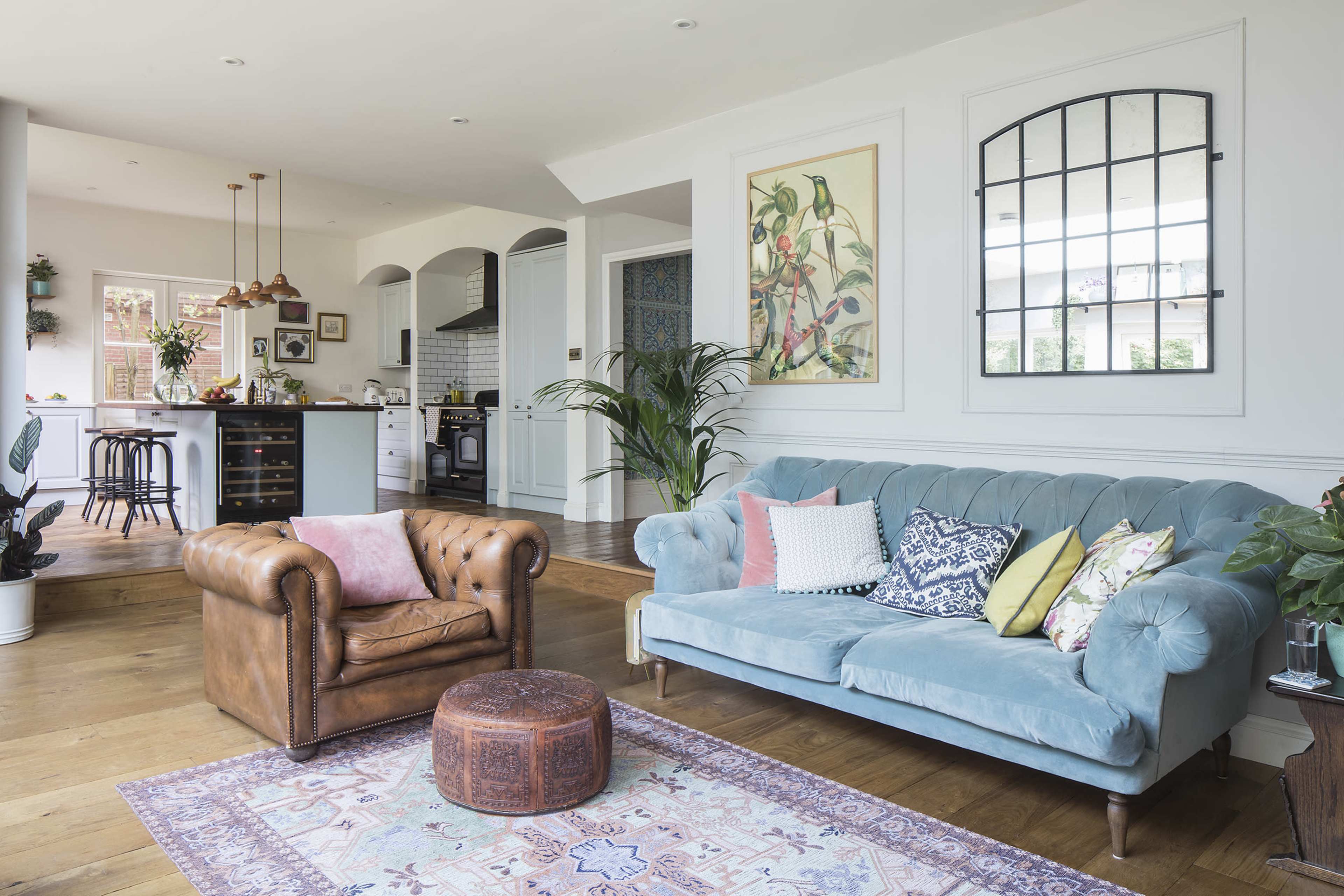 A cozy living room featuring a blue velvet sofa, a brown leather armchair, and a patterned rug, with a kitchen visible in the background.