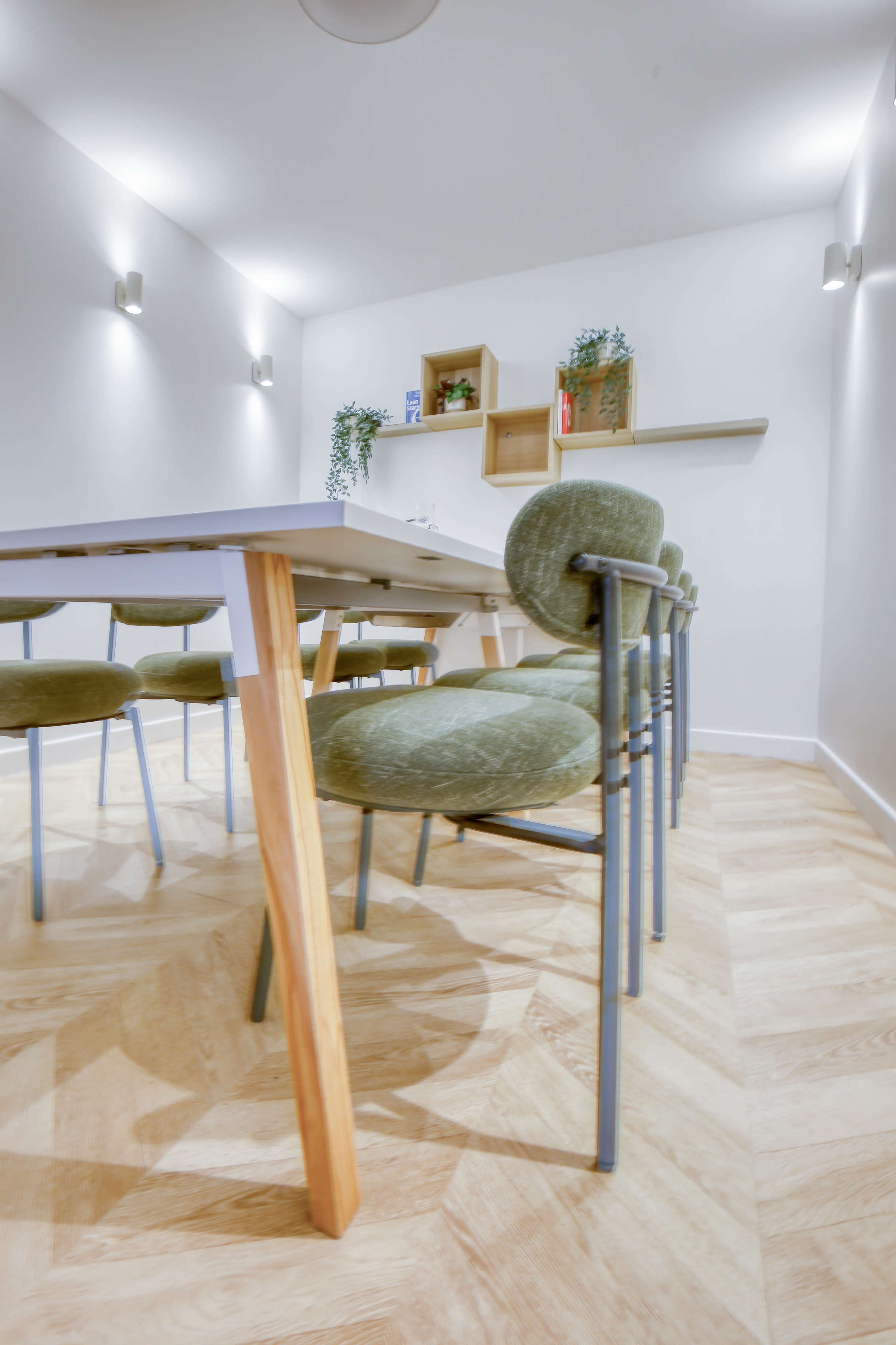 The image shows a modern meeting room with a long table and rows of green chairs set on a light wooden floor.