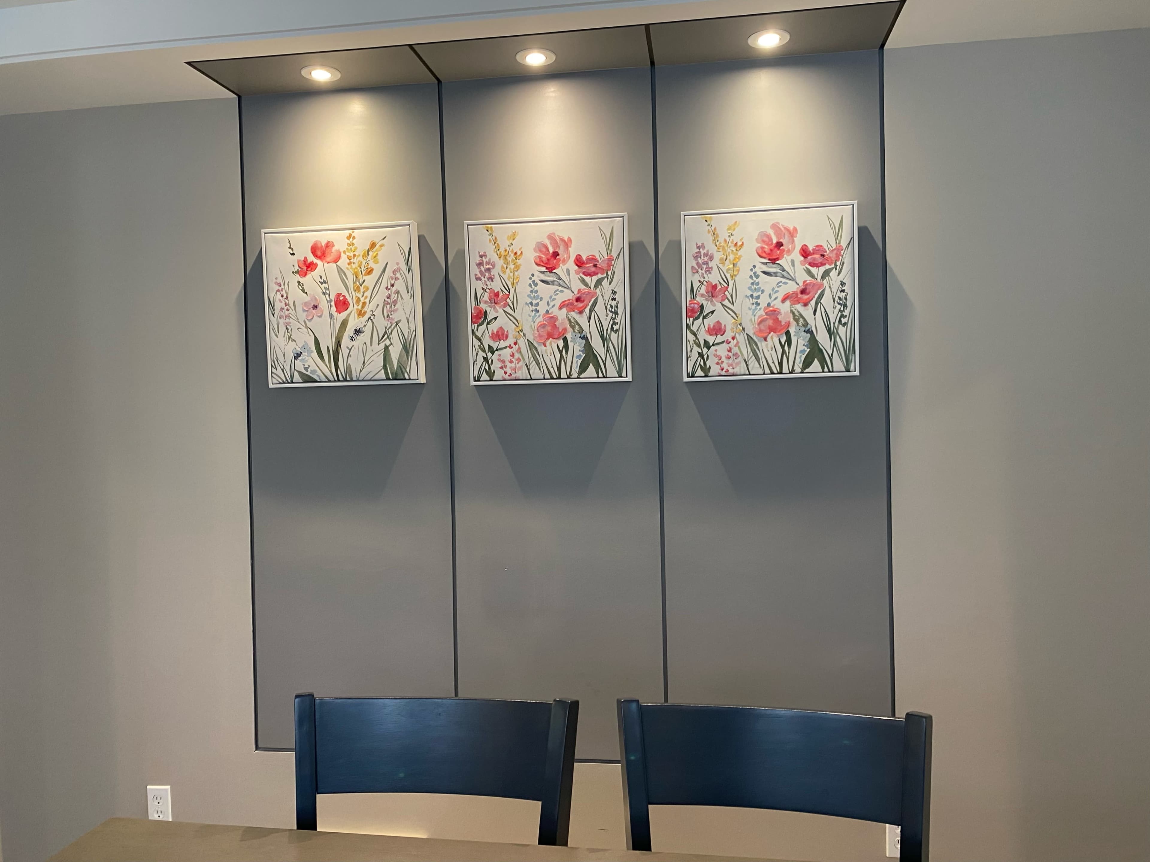 A dining area featuring a gray wall with three framed floral paintings and two blue chairs at a table.
