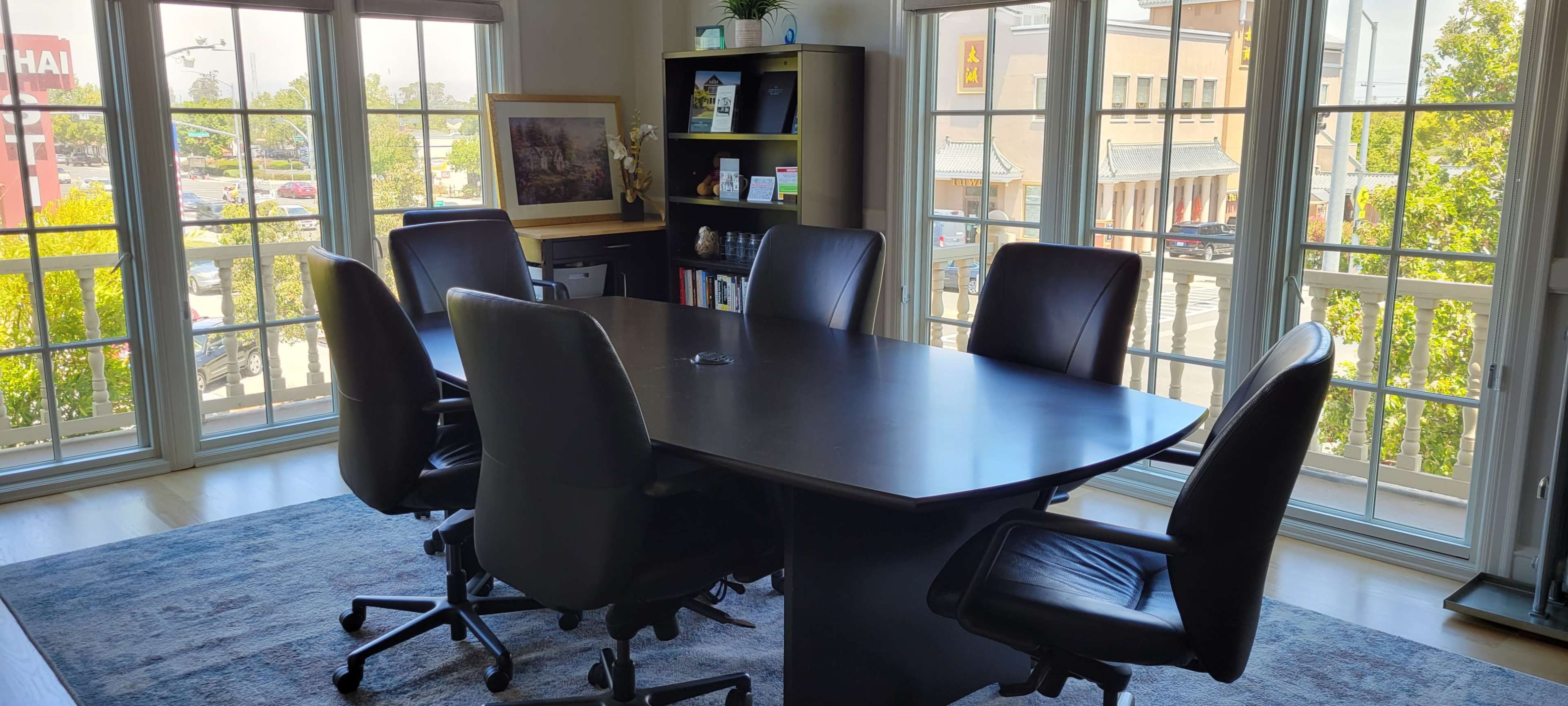 A conference room features a large oval table surrounded by six black leather chairs, with a view of a busy street through large windows.