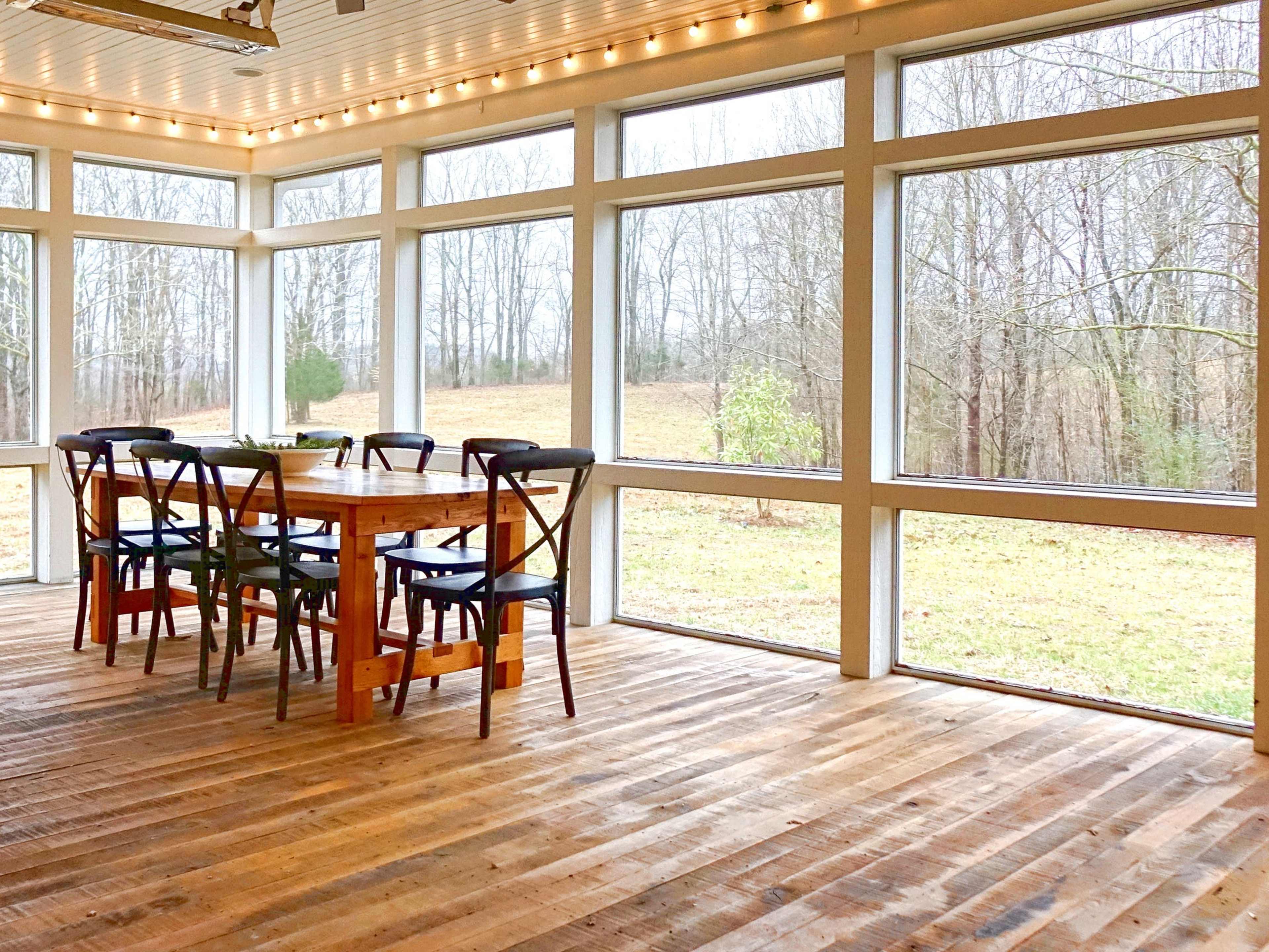 A spacious room features a wooden dining table surrounded by chairs, with large windows offering a view of a grassy area and trees outside.