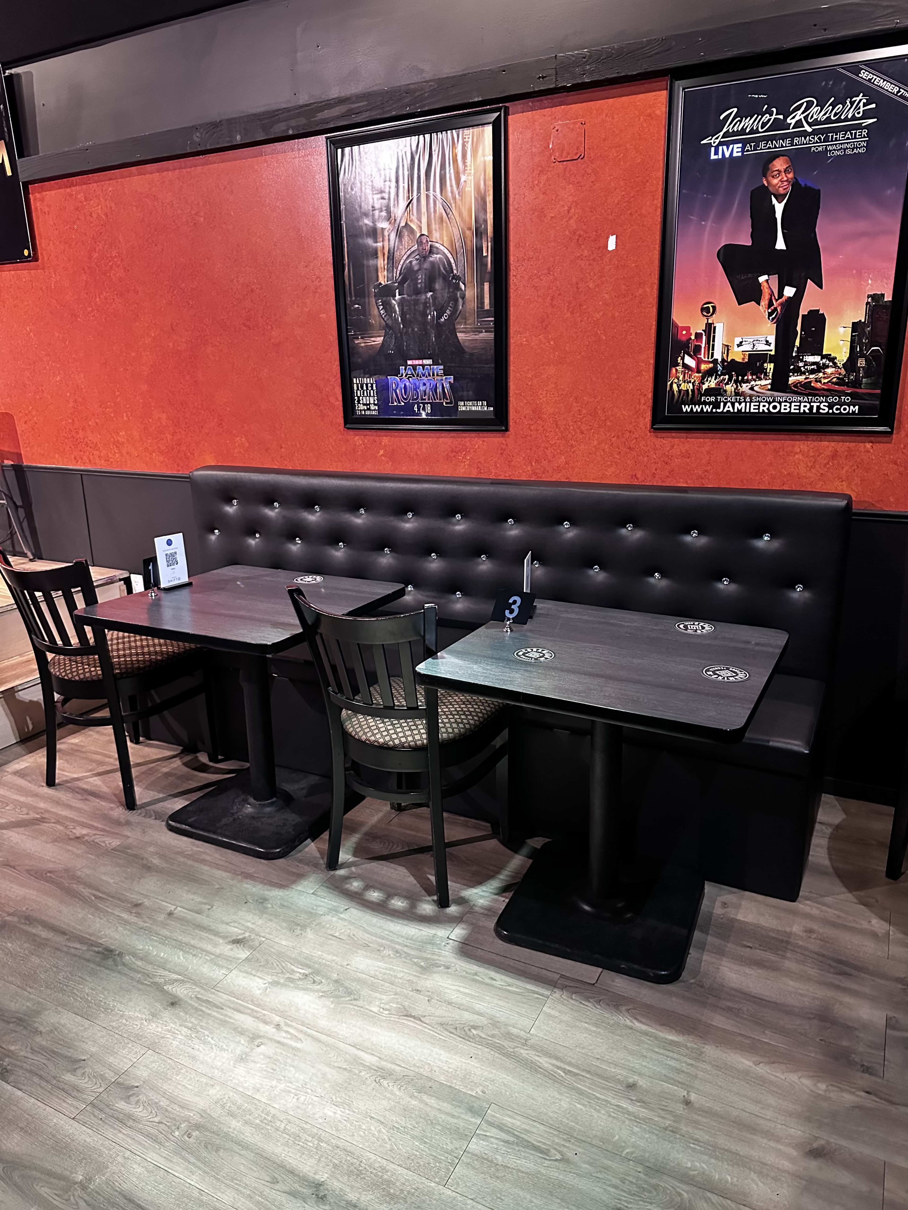 Two empty tables with black chairs in a restaurant, set against a vibrant orange wall adorned with a large framed poster.