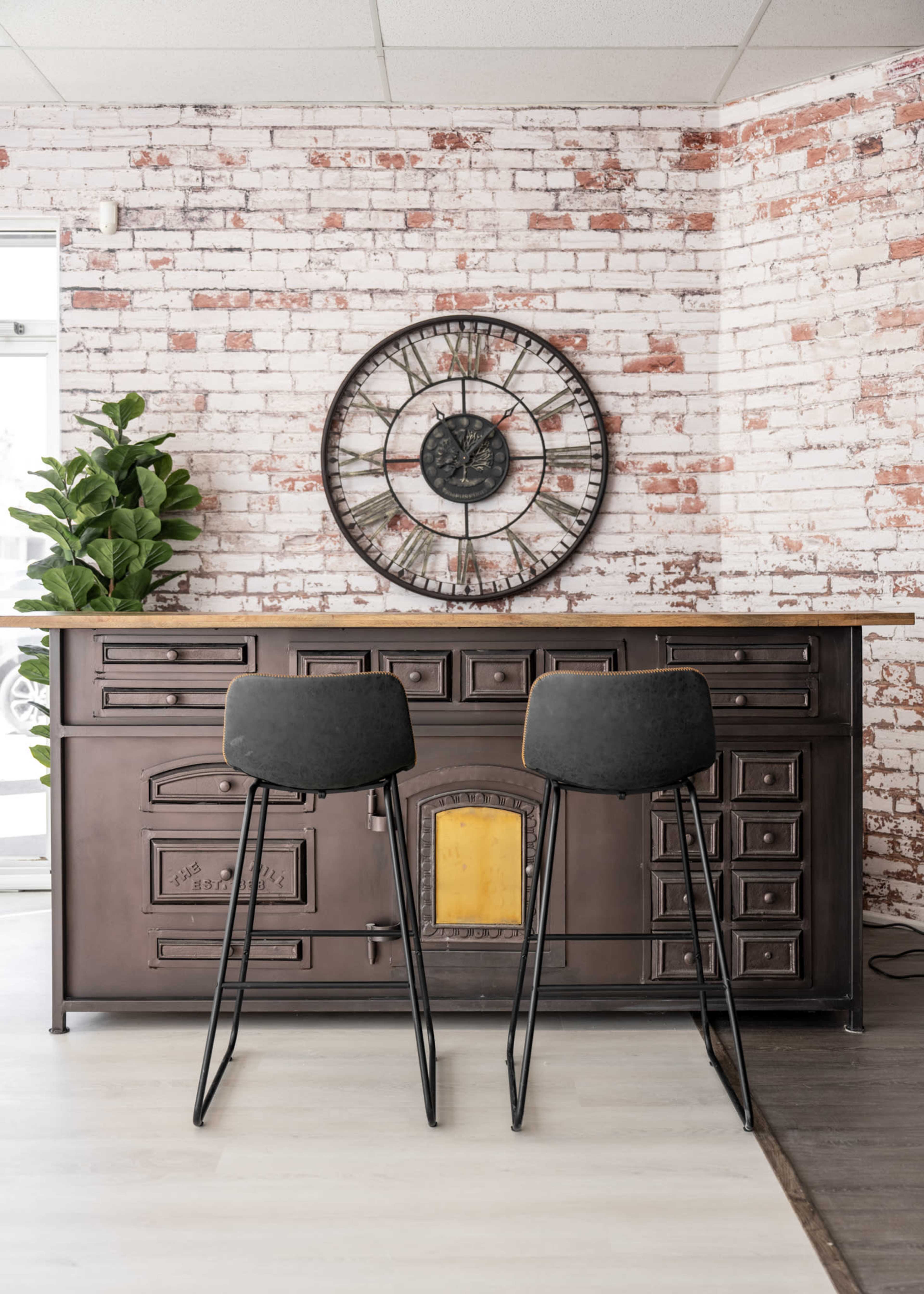 A rustic kitchen space features a metal bar counter with a yellow accent and two black stools, against a background of exposed brick walls and a large wall clock.