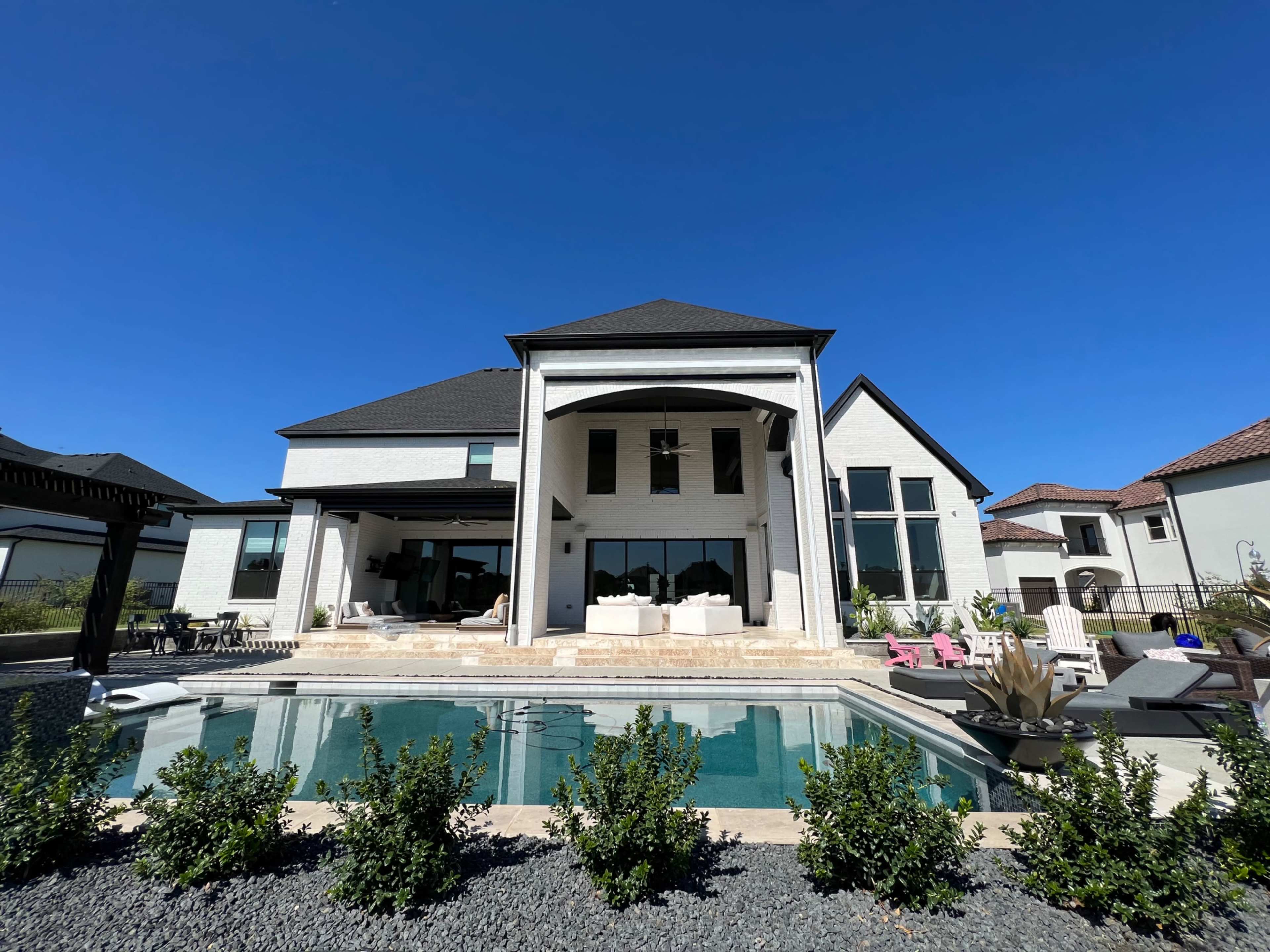 The image shows a contemporary two-story house with large windows and a pool in the foreground, surrounded by low shrubs under a clear blue sky.