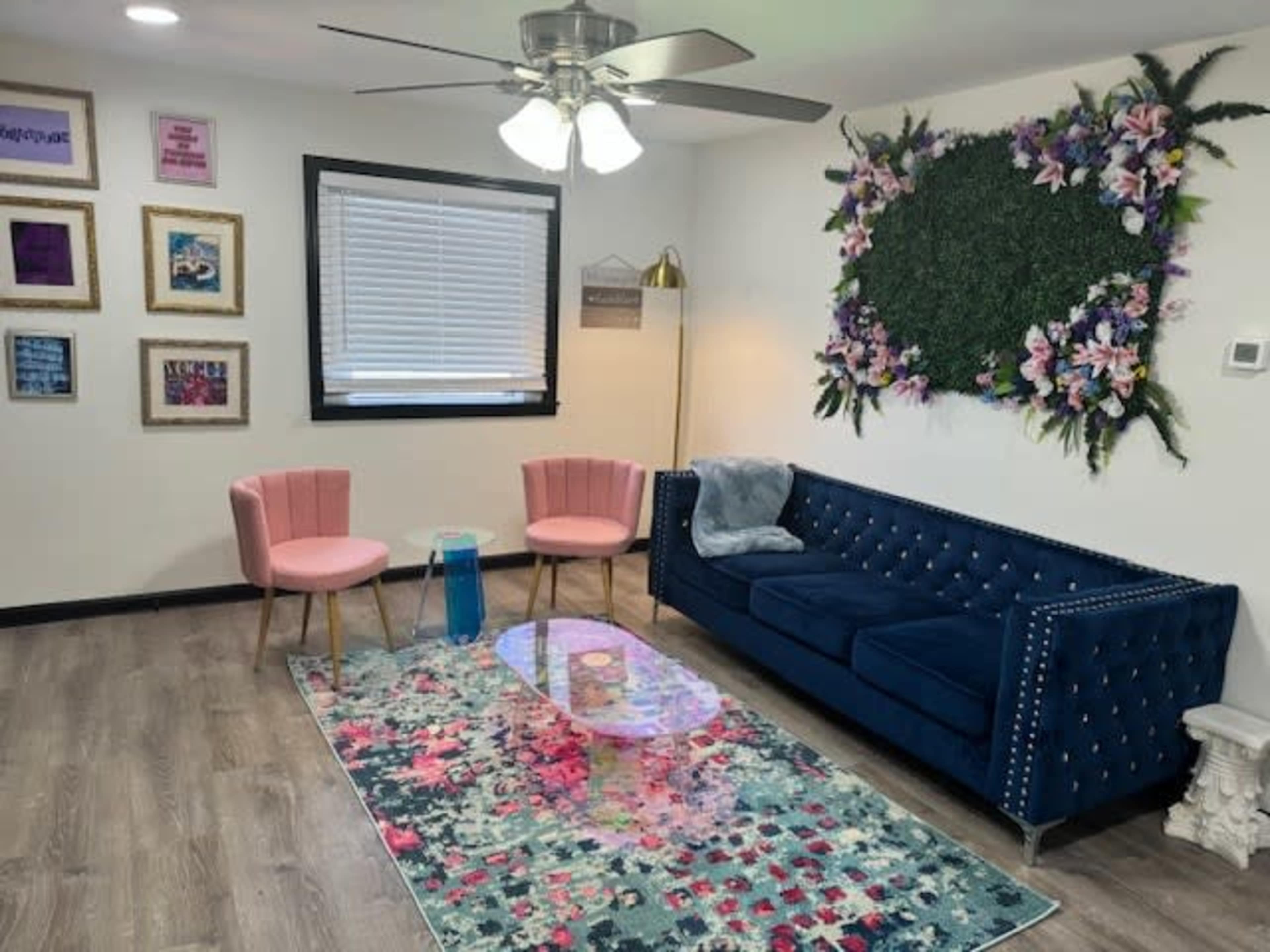 The room features a blue velvet sofa, two pink chairs, a colorful area rug, and a wall adorned with framed artwork and a floral design.