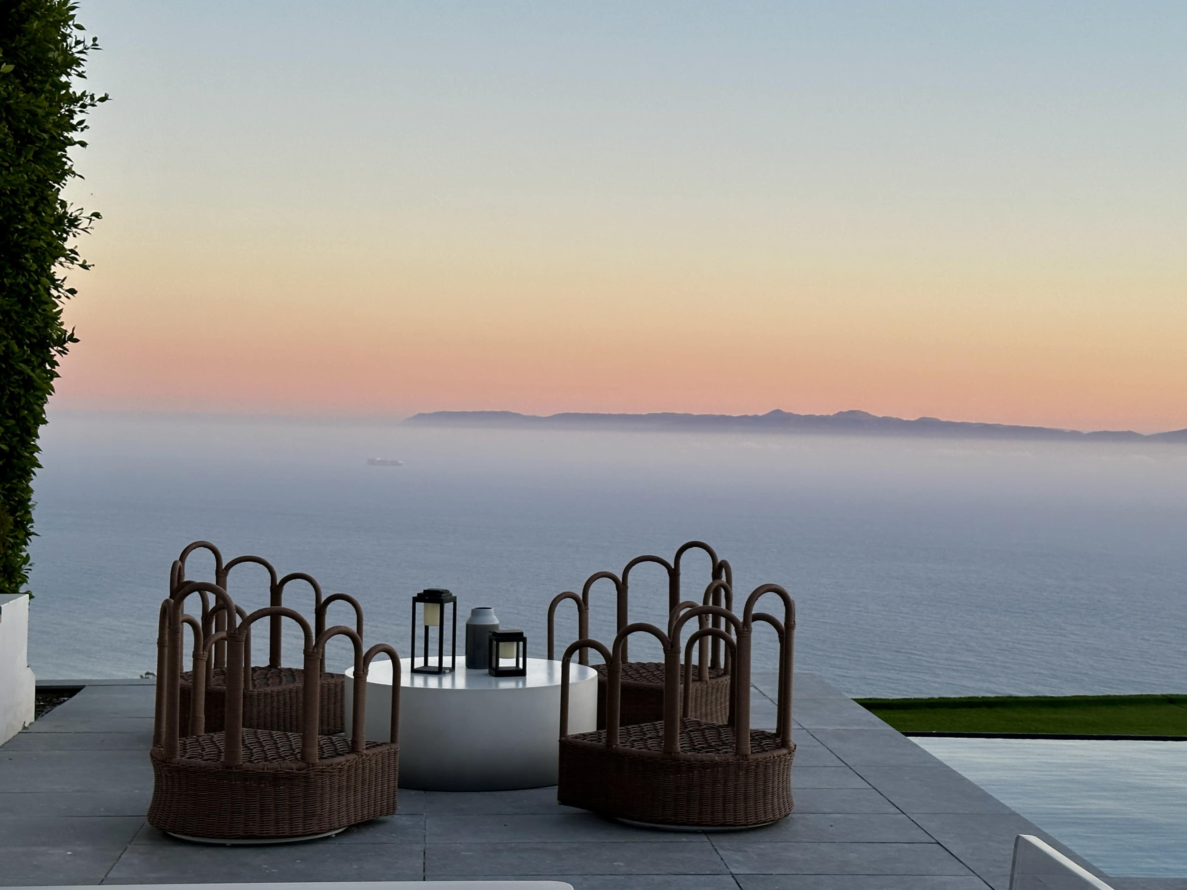 A terrace features two woven chairs and a circular table, overlooking a misty ocean with a sunset sky.